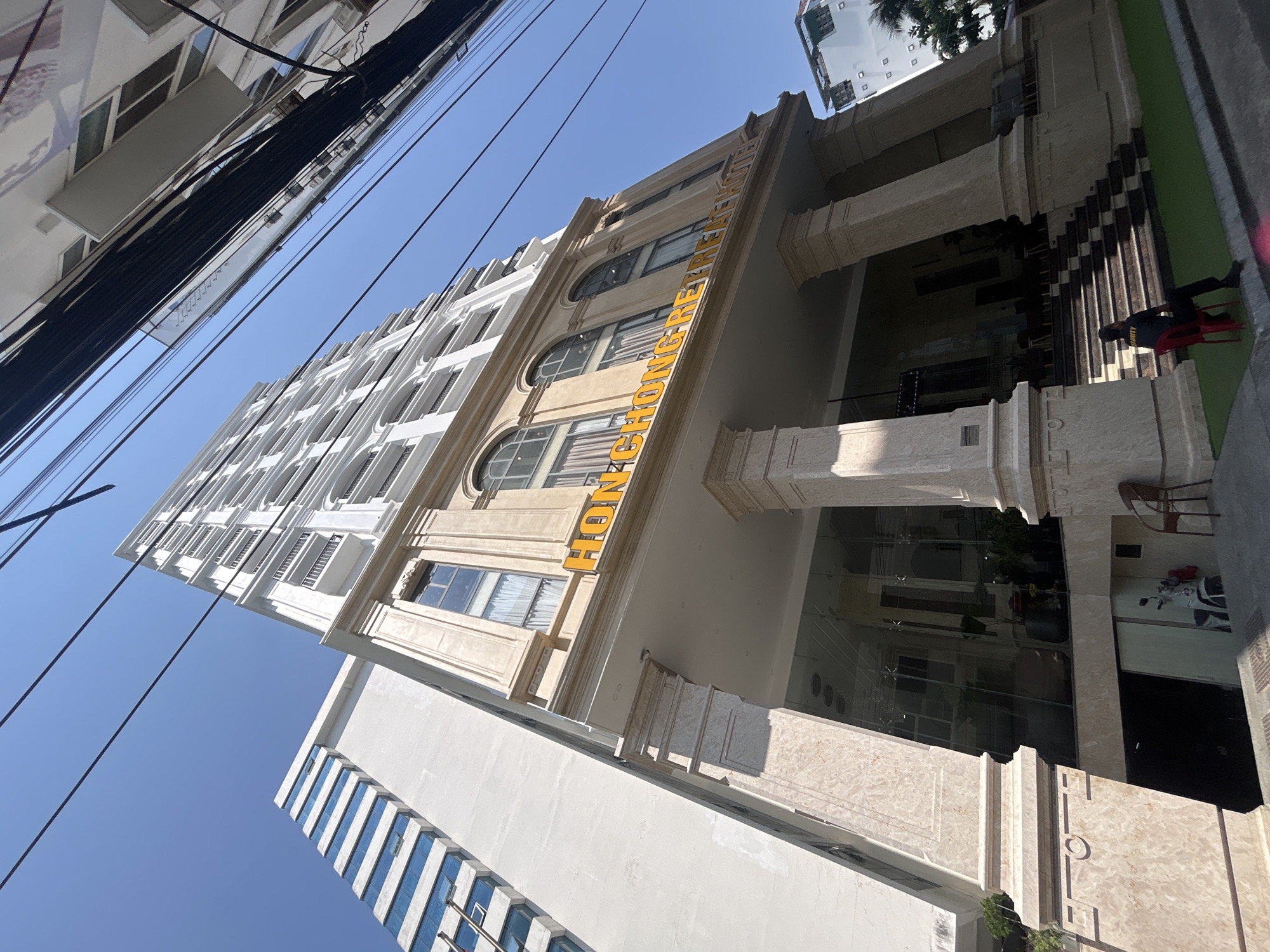 An upward-angled exterior shot captures two buildings in Bắc Nha Trang, Vietnam, on a sunny day. The building on the left is mostly white with repeating horizontal windows. The building on the right, likely a hotel, is partially covered in yellow signage reading "HON CHONG RETREAT." The entrance features marble pillars, glass walls, and a few plants. A security guard sits in a red chair at the entrance, next to an artificial turf area. Overhead are multiple black electrical or utility wires against a clear blue sky. The image's perspective makes it difficult to ascertain the exact number of floors of the buildings.