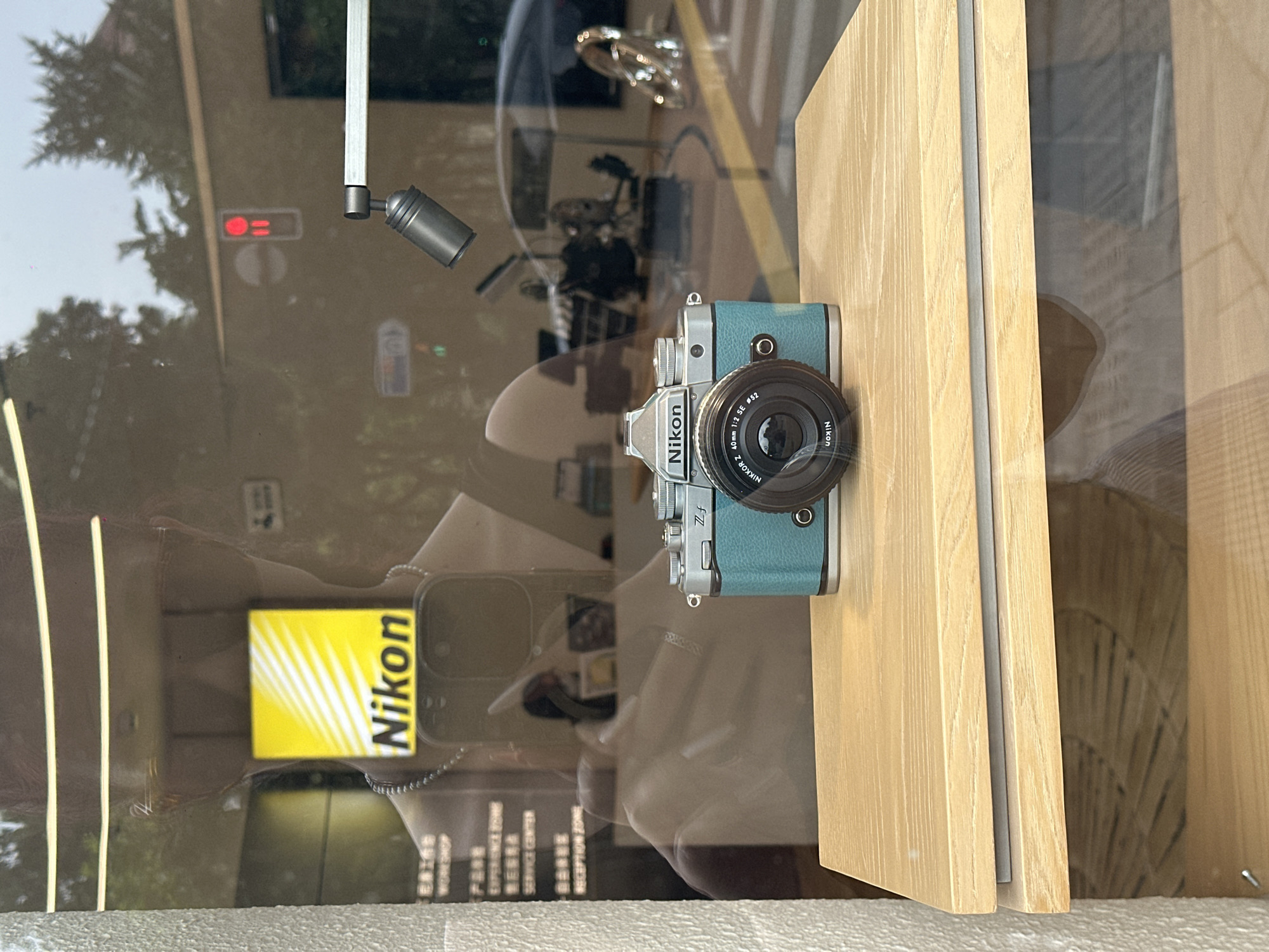 This image depicts a Nikon Zf camera displayed on a light wooden shelf, likely in a retail setting or showroom. The camera is teal and silver with a black lens. Behind the camera, a glass window reveals reflections of the surrounding environment. The reflections show a person, a yellow and white Nikon logo, trees, and buildings, suggesting an outdoor urban setting. Notably, the reflection of a red traffic signal is visible. The text on the glass also indicates it is in Shanghai, China, with some text visible in Chinese characters, possibly referring to a service center or experience zone. The overall scene is a well-lit indoor space with a focus on the camera, framed by the reflections in the glass.