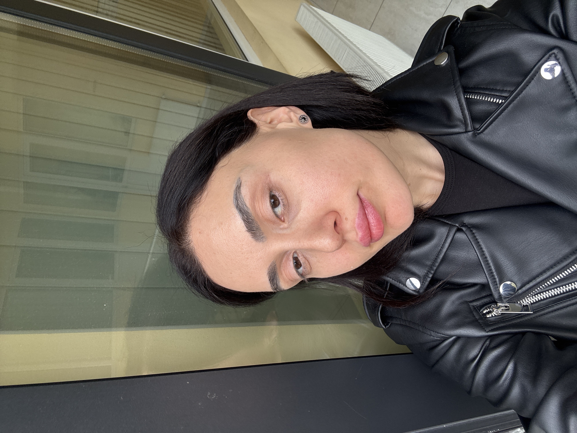 A woman is facing the camera, wearing a black leather jacket and a black shirt. She has dark hair and light skin. The background shows a window and part of a building. The exact location in Tbilisi, Georgia, is not discernible from the image.