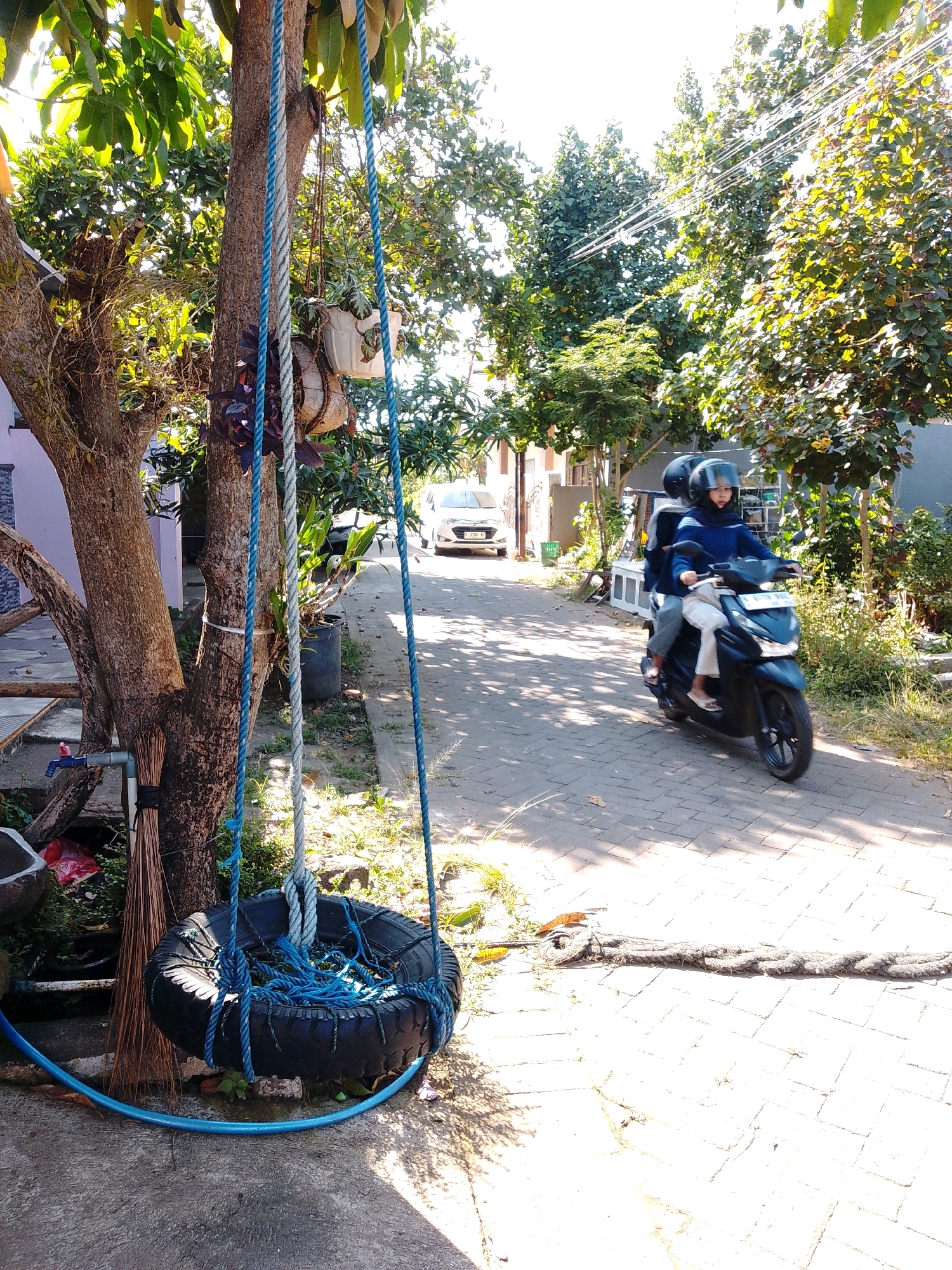 In the city of Cikal, Indonesia, a sunny day illuminates a residential street paved with interlocking bricks. On the left side of the frame, a tire swing hangs from a tree, its blue ropes taut. To the right of the tree, a broom leans against its trunk, and a blue hose snakes across the ground. Further down the street, two people on a scooter, both wearing helmets and face coverings, ride away from the camera. The scooter, a dark color, is moving. In the background, a white car is parked, and lush green trees and vegetation line the street, casting dappled shadows. The scene captures a moment of everyday life on a warm Indonesian day.