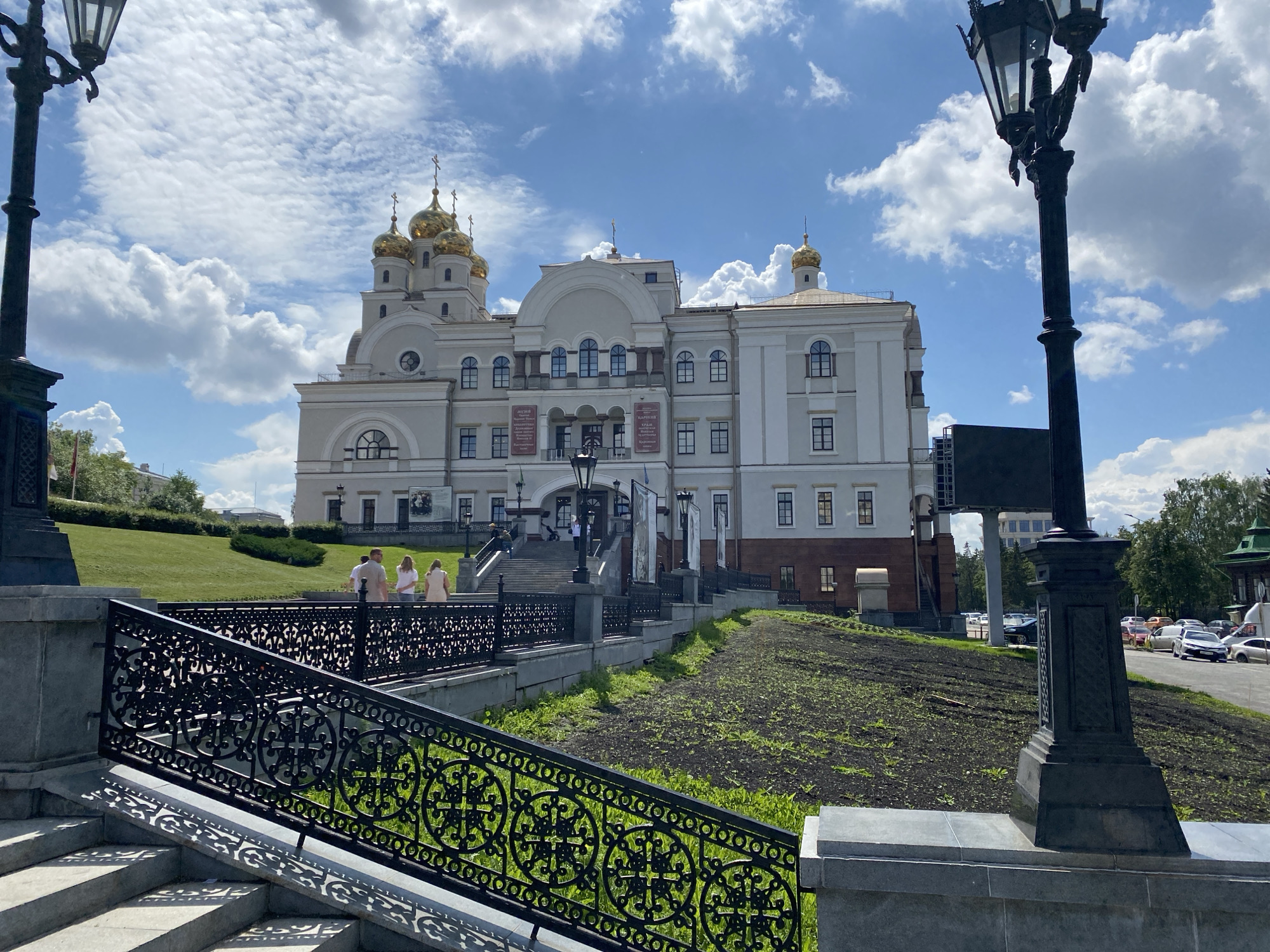 The image depicts the Holy Trinity Cathedral in Yekaterinburg, Russia, on a bright, partly cloudy day. The grand, white-and-gray cathedral features multiple golden domes topped with crosses, and its facade is adorned with arches and rectangular windows. A wide, stone staircase leads up to the entrance, flanked by ornate black metal railings.
In the foreground, a large black lamppost stands to the right, its detailed design visible against the bright sky. To the left, another lamppost, partially visible, frames the scene. Several people are visible on the walkway leading to the cathedral stairs, some walking and others standing. The area around the cathedral is landscaped with green lawns and dark soil beds, some appearing freshly tilled. A street with parked cars is visible in the background, indicating an urban setting. The overall impression is one of a prominent religious and civic landmark bathed in sunlight.