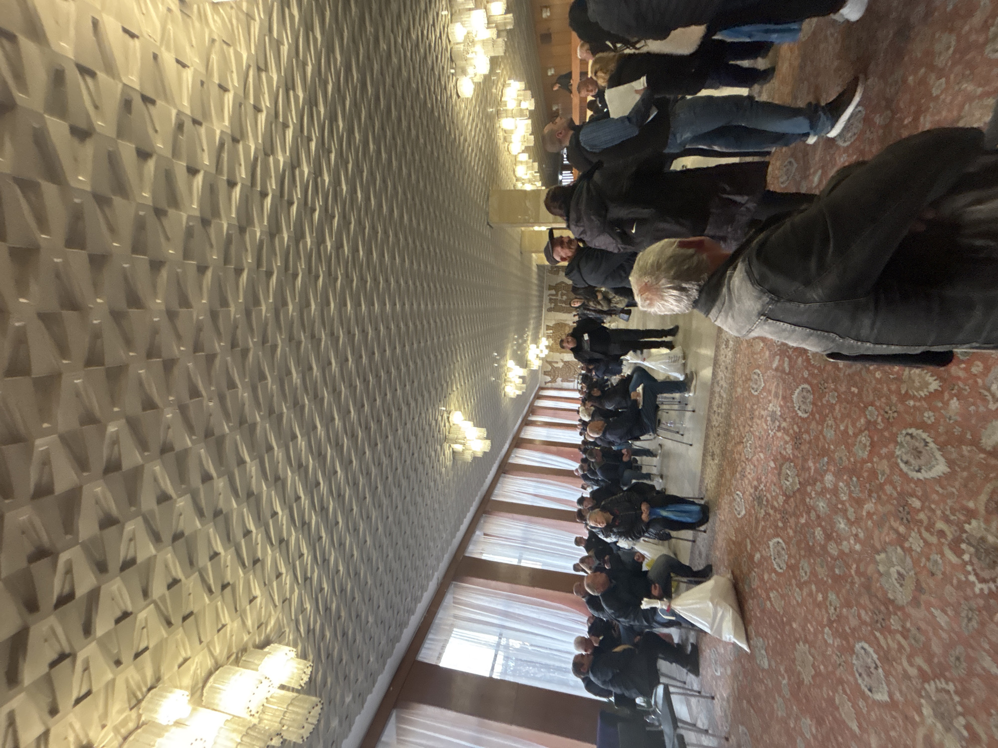 A long, brightly lit hall with textured white walls and ornate chandeliers houses a gathering of people. Many individuals are standing while others are seated in rows of chairs. The floor is covered with a patterned carpet, and there are windows with white curtains visible along one side of the hall. The location is consistent with an event or assembly taking place in a large public space, such as a municipal hall in the city of Kardzhali, Bulgaria.