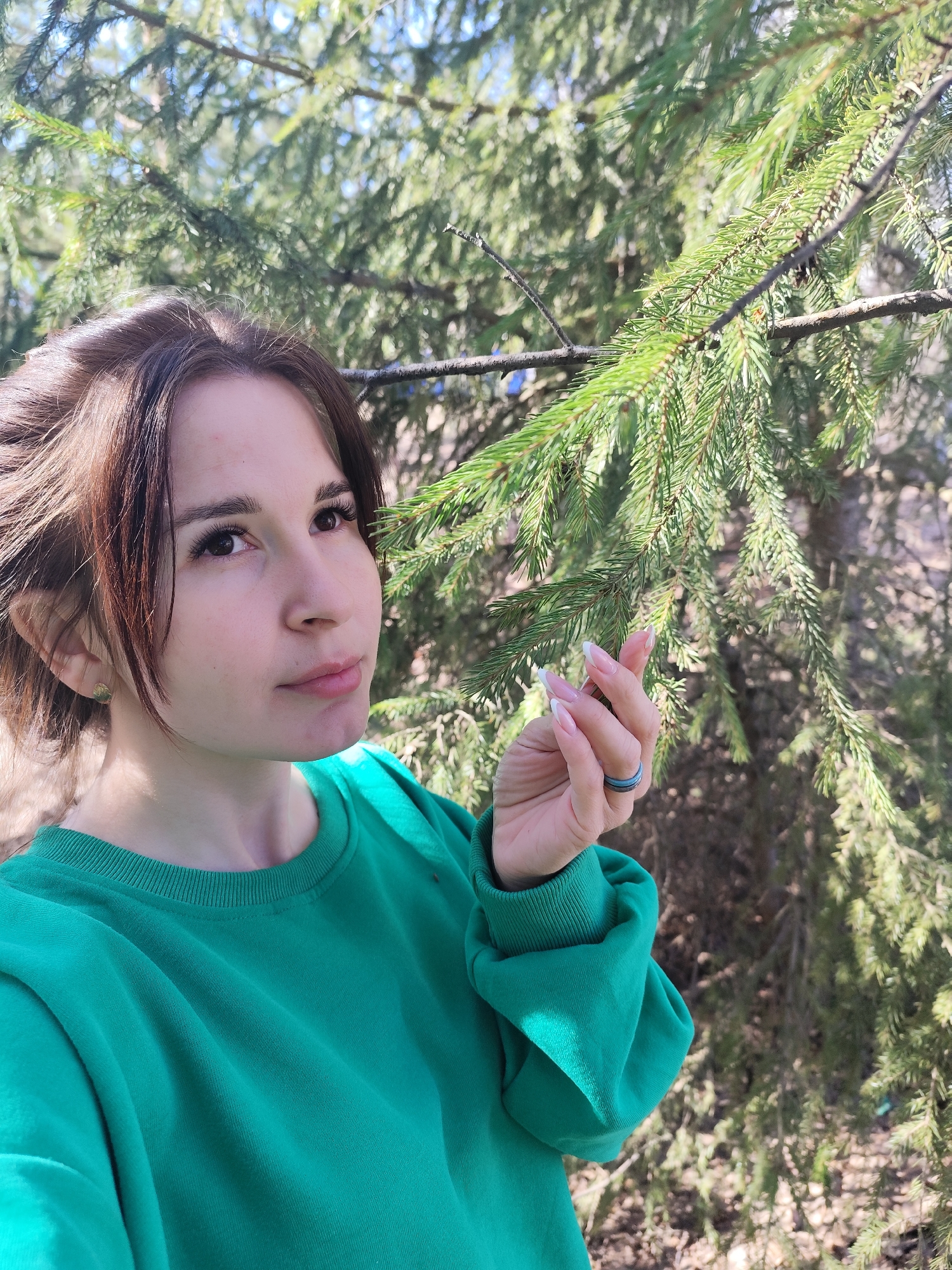 A young adult woman is pictured outdoors in a sunlit coniferous forest, likely in Фоки, Russia. She is seen from the upper chest up, angled slightly to her right, and looking upwards and to her right with a calm, reflective expression. Her dark brown hair is styled back, with some wisps framing her face, and she has light skin, dark eyes, and full lips.
She is wearing a vibrant, possibly emerald green, long-sleeved sweatshirt. Her left hand, adorned with manicured nails featuring french tips and a blue/teal ring on her ring finger, is gently reaching towards or lightly touching a branch of a pine or spruce tree. A small, golden-toned earring is visible in her left ear.
The background is dominated by the lush green needles and branches of numerous coniferous trees, indicating a natural, wooded setting. The bright sunlight creates clear highlights on the tree foliage and the woman's hair, suggesting a clear day, possibly mid-morning or afternoon. The ground in the distance is sparsely covered with dry leaves or pine needles. The scene conveys a tranquil moment amidst nature. No text is visible in the image.