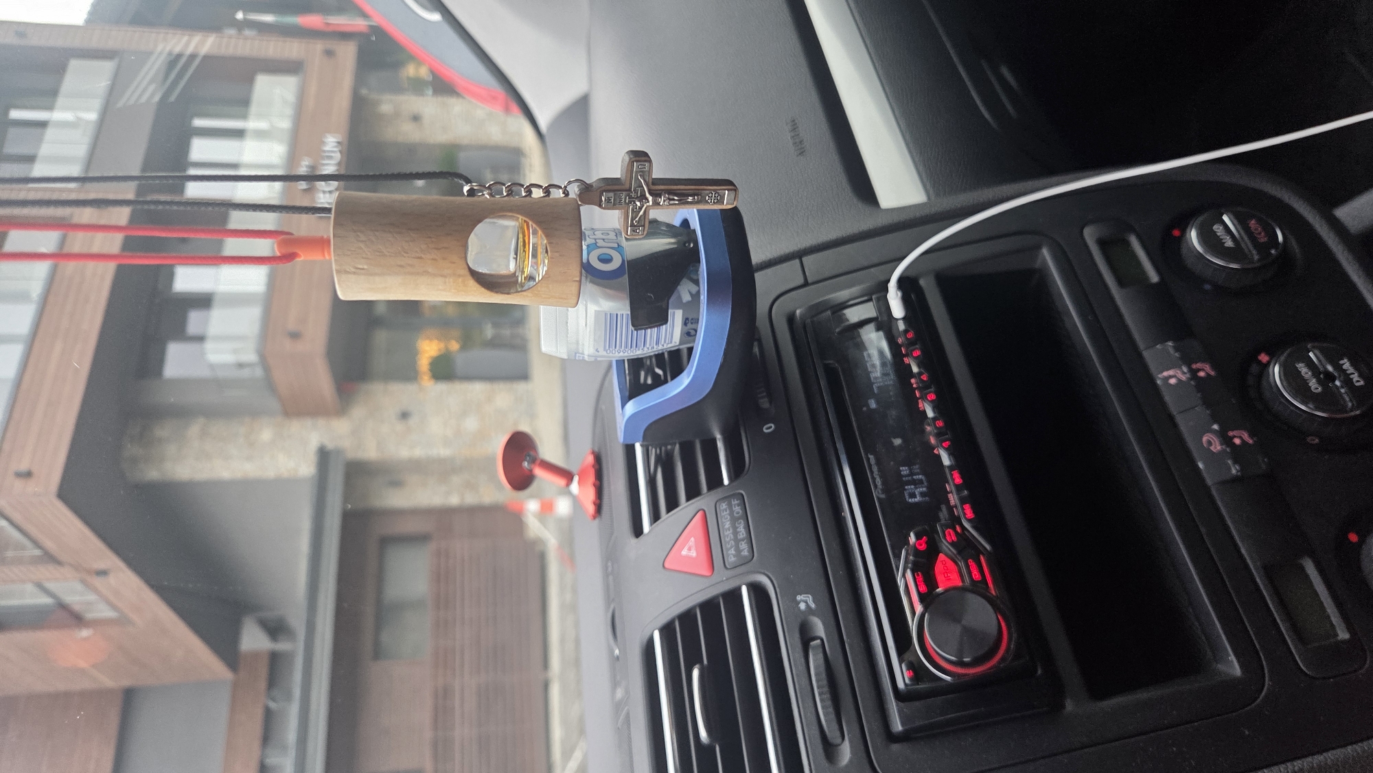 The image shows the interior of a car, specifically the dashboard and a section of the windshield. A wooden car air freshener with a glass vial containing liquid and a crucifix charm hangs from the ceiling. In the foreground, a car stereo with a red illuminated display showing "AUX" is visible. To the left of the stereo, air vents and a "PASSENGER AIR BAG OFF" indicator are present. In the background, through the windshield, a modern building with wooden and stone elements is visible. The setting appears to be outdoors, given the view through the windshield.