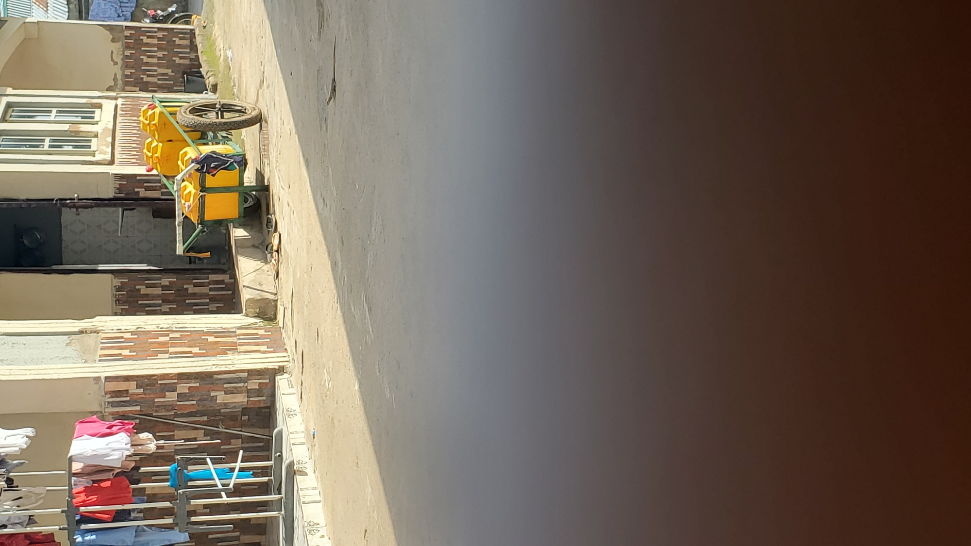 This media file depicts an outdoor scene in Jalingo, Nigeria. The image is oriented vertically. The foreground shows a clothes drying rack with several items of clothing, including pink, white, red, and blue garments, neatly folded or hung. Beyond the drying rack, there is a textured wall with horizontal bands of brown and beige tiles.
Further in the background, a yellow cart with several large yellow containers and a wheel is visible, possibly used for transporting water or other goods. This cart is positioned on a paved surface. To the left of the cart are windows of a building, some with multiple panes and white frames. The building features patterned tiles in a similar style to the wall in the foreground. A dark doorway or opening is also present. In the upper left corner, a glimpse of a motorcycle is visible, suggesting activity in the vicinity.
The scene appears to be set during the daytime, with bright sunlight casting shadows, indicating clear weather. The overall impression is one of a residential or commercial area, with everyday activities like laundry taking place. There is no visible text in the media file. The image is slightly tilted, and a large portion of the right side is blurred and dark, obscuring details in that area.