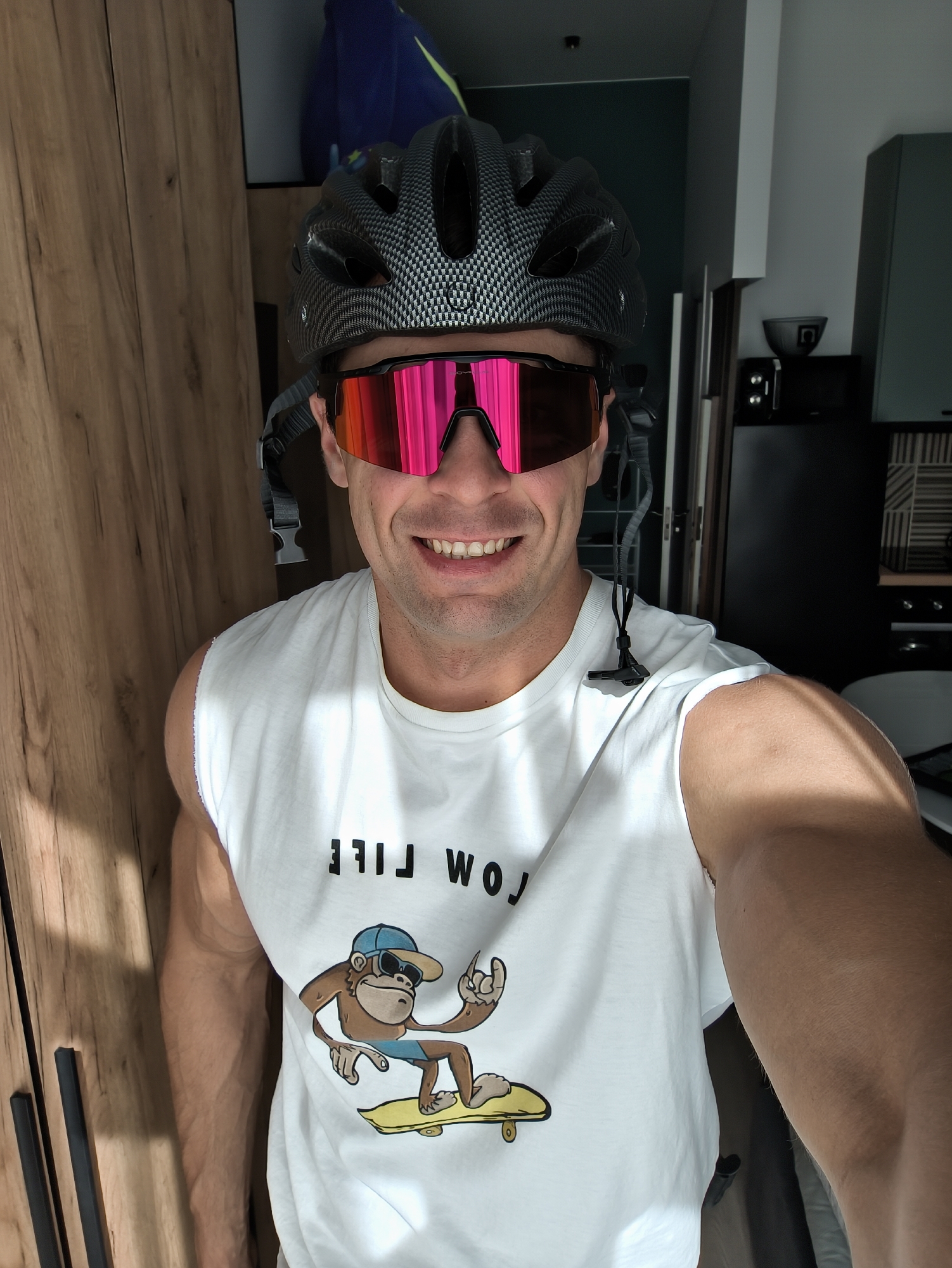 A male subject is captured in a front-facing selfie, positioned indoors. He appears to be Caucasian, with a muscular build, smiling broadly with visible teeth. He wears a dark gray cycling helmet with a carbon fiber pattern and large, reflective pink sport sunglasses. A white, sleeveless t-shirt, possibly a cut-off, features inverted black text that reads "LOW LIFE," beneath which is a cartoon depiction of a monkey wearing a blue baseball cap and sunglasses, in blue swim shorts, riding a yellow skateboard. The monkey's right hand forms a "rock on" gesture. A black lanyard or strap with a small black attachment hangs from the right side of his head/helmet.
The background to the left consists of a light brown wooden wardrobe or cabinet with vertical black handle strips. Above the wardrobe, a glimpse of a blue fabric item with a yellow-green accent is visible. To the right, the background reveals an interior space, likely a kitchen, with dark gray or black cabinetry. A small black appliance, possibly a microwave or mini-oven, with illuminated controls, sits on a counter, with a dark bowl or container above it. The lighting suggests an indoor setting, possibly with ambient light from the right, casting a shadow on the subject's right arm. The overall environment appears to be a modern residential apartment.