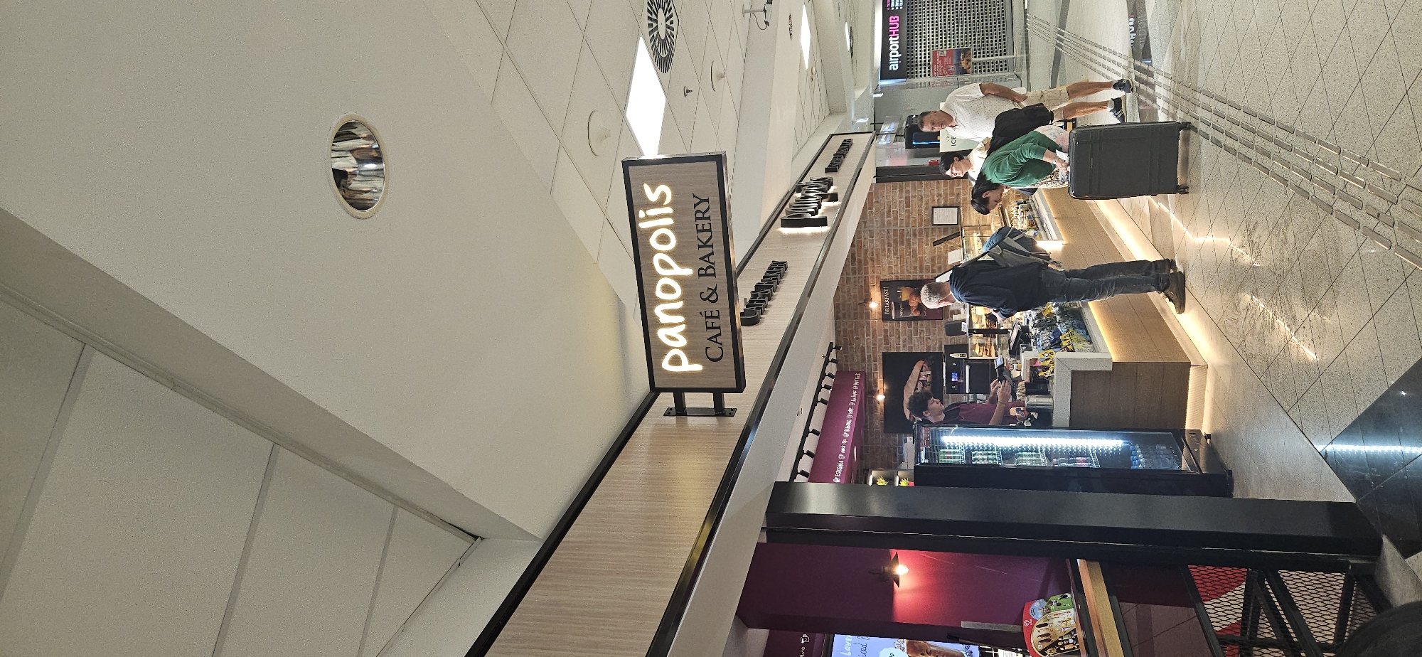The image shows a busy airport terminal at Budapest Ferenc Liszt International Airport (BUD) in Budapest, Hungary. A cafe and bakery called "Panopolis" is featured prominently, with customers ordering and waiting for their food. The cafe has a modern design with wooden accents and bright lighting. People of various ages are seen in the scene, some with luggage, indicating travel. The floor is tiled and reflective, with tactile paving indicating a pathway. A row of rolling shutters for shops is visible in the background, along with airport signage. The overall atmosphere suggests a typical airport environment with commercial activity.