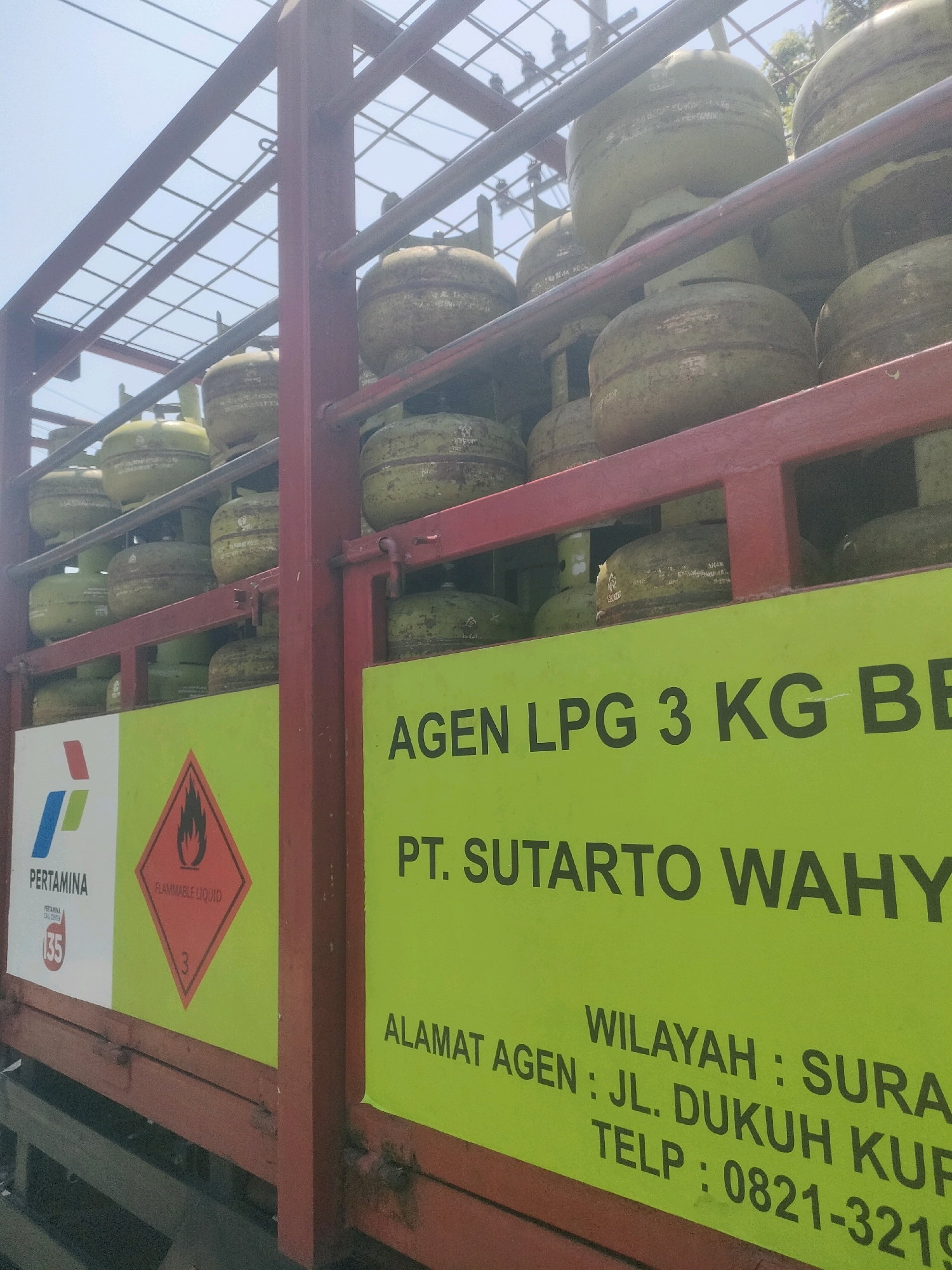 A truck bed is loaded with many green propane tanks. The truck bed has a red metal frame. A large green sign is attached to the side of the truck bed with text printed in black. The text reads "AGEN LPG 3 KG BE PT. SUTARTO WAHYU WILAYAH: SURA ALAMAT AGEN: JL. DUKUH KUP TELP: 0821-3219". To the left of this sign is a white sign with the Pertamina logo and a red diamond-shaped hazard symbol indicating "FLAMMABLE LIQUID". The sky is visible above the truck bed, which is full of propane tanks. This image was taken in Surabaya, Indonesia.