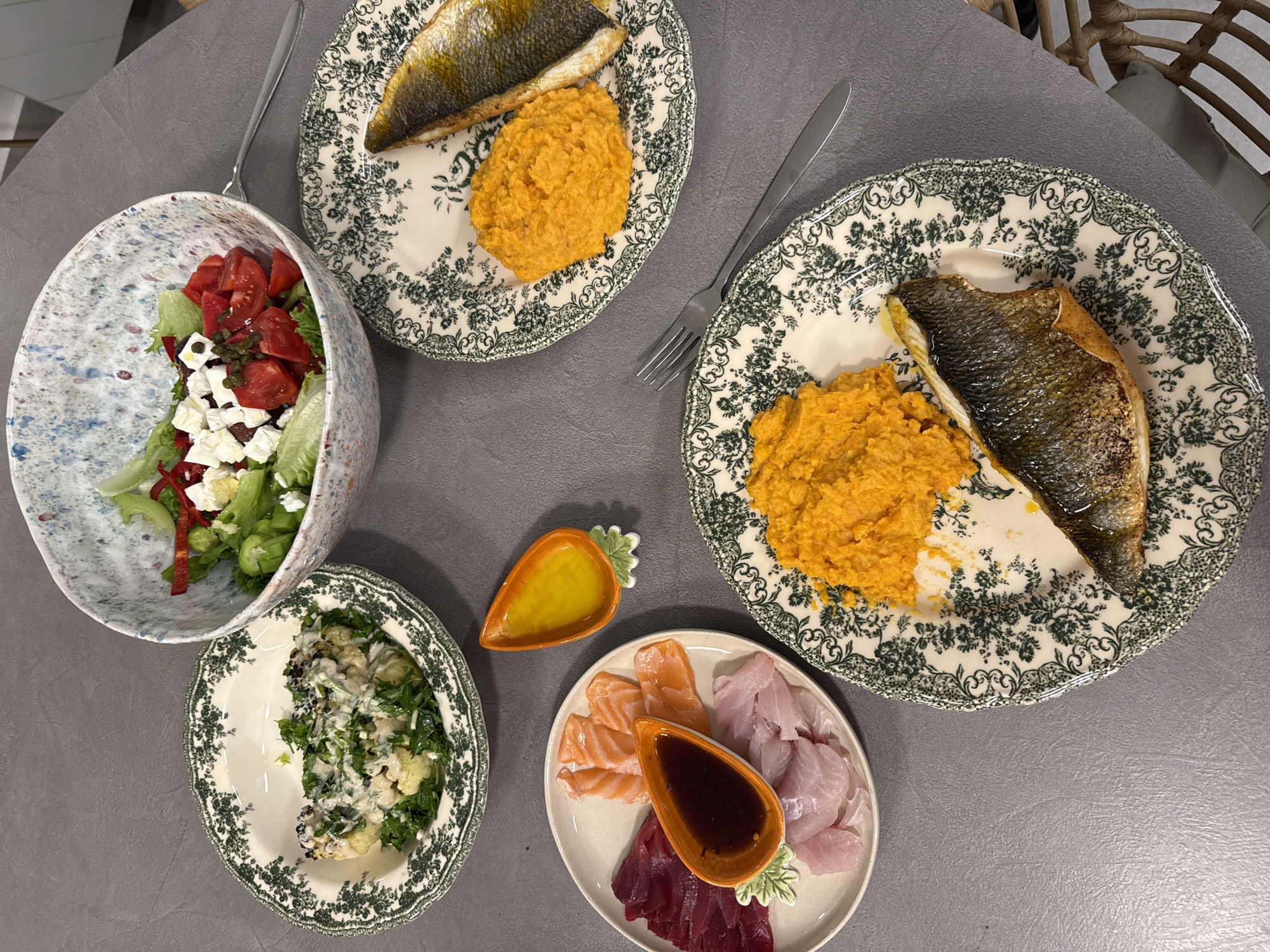The image shows a table setting with several plates and bowls of food, suggestive of a meal. There are two plates with patterned rims, each containing a piece of fish and a portion of mashed orange vegetable, likely sweet potato or squash. A bowl holds a mixed salad with lettuce, tomatoes, peppers, and white cheese. Another plate has what appears to be cauliflower with a white sauce. A final plate displays an assortment of raw fish, possibly sashimi, along with two small containers holding different sauces. The setting appears to be indoors, possibly a home or restaurant. There is no indication of the location being Tel-Aviv, Israel in the image.
