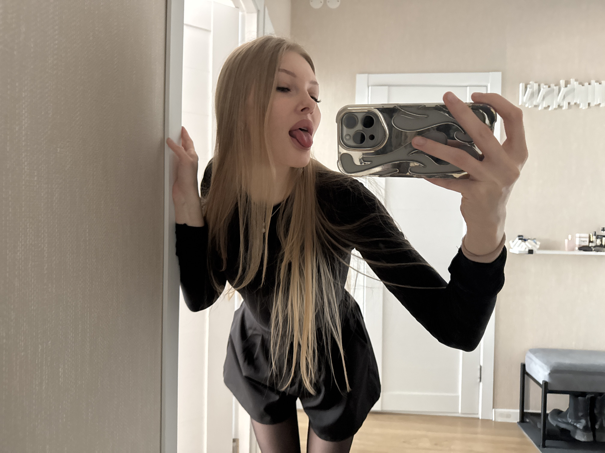 The image depicts an interior residential scene in Perm, Russia. A female subject with long, straight blonde hair is oriented frontally but angled slightly right, appearing to take a self-portrait using a smartphone reflection. Her head is tilted upwards, with closed or squinted eyes and an extended tongue. She is dressed in a black long-sleeved top, black shorts or a romper, and black hosiery. Her left hand holds a smartphone encased in a grey, relief-patterned cover, displaying a visible triple-lens camera array. Her right hand makes contact with a white door frame on the left side of the frame. The background features a light beige textured wall on the left and a smooth light beige wall on the right. A white, multi-segment horizontal wall light fixture is mounted on the right wall. Further right, a white floating shelf holds assorted small objects, including cosmetic items. Below the shelf, a grey upholstered bench with black metal legs is positioned, with black boots resting on a black mat beneath it. The flooring is light brown wood. The environment is consistent with a modern residential hallway or entryway.