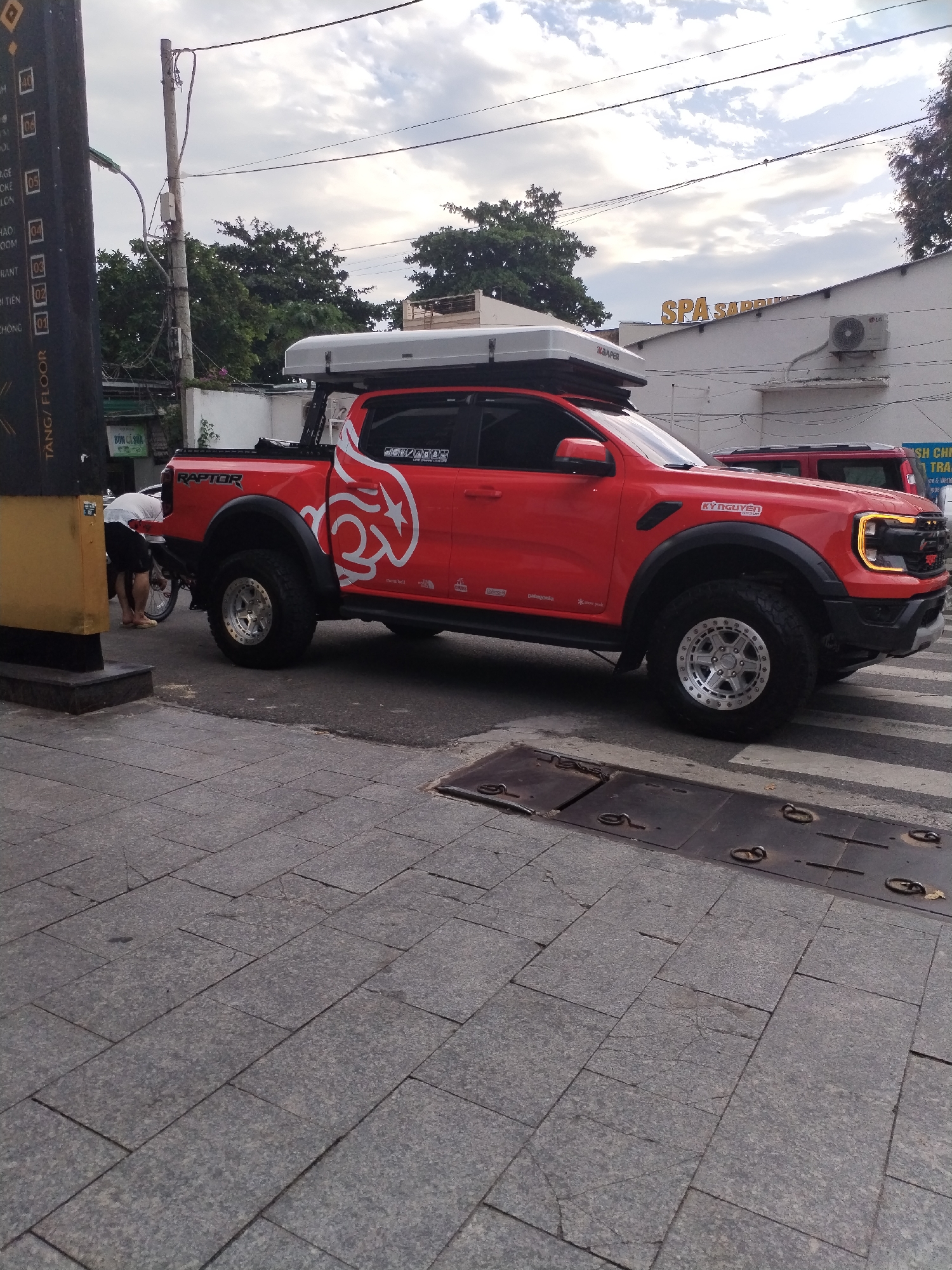 A customized red Ford Ranger Raptor pickup truck, equipped with a roof-top tent branded "KAMPER," is parked on a city street next to a sidewalk. The truck features custom wheels and tires, "RAPTOR" branding, and decals including "KỲ NGUYÊN," "mont bell," and "patagonia." The sky above is overcast, suggesting daytime with diffuse light.
To the left of the truck, a person is bent over a scooter or motorcycle. A tall, black vertical sign is visible on the left, displaying floor numbers (01-08) and text like "TANG/FLOOR," "BUN CÁ SỬA," "HAO OOM," "DI TIÊN," and "CHÔNG," indicating a multi-story building with various establishments. In the background, a building features large golden letters spelling "SPA SAPP" (likely "SPA SAPPHIRE") and an LG air conditioning unit. Another red vehicle is partially visible behind the main truck.
The ground consists of grey rectangular paving tiles on the sidewalk and asphalt with a white crosswalk on the street. A utility pole with numerous wires is present in the mid-ground. The visible Vietnamese text and urban setting, consistent with Bac Nha Trang Ward, Vietnam, provide strong location cues.