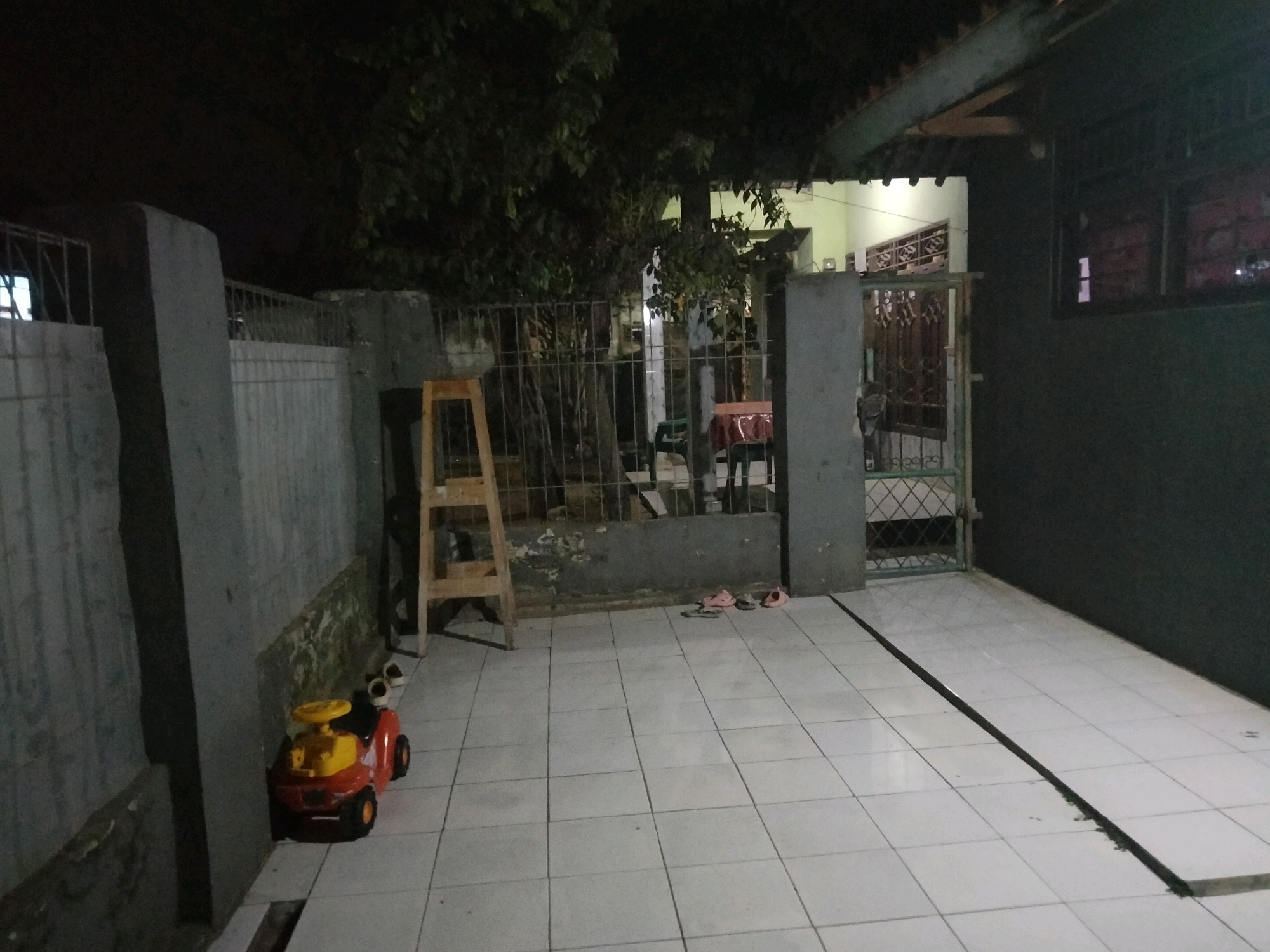 The image depicts an outdoor scene at night, likely a residential area in Suka Sari, Indonesia. The foreground is a tiled patio area with a black drain cover running diagonally across it. To the left, a red and yellow toy car is parked near a grey concrete wall. Further back, a wooden stepladder stands in front of a metal fence. Beyond the fence, trees and foliage are visible, with a building in the background illuminated by interior lights. Two pairs of pink sandals are placed on the patio near the fence. The overall impression is of a somewhat cluttered residential yard at night.