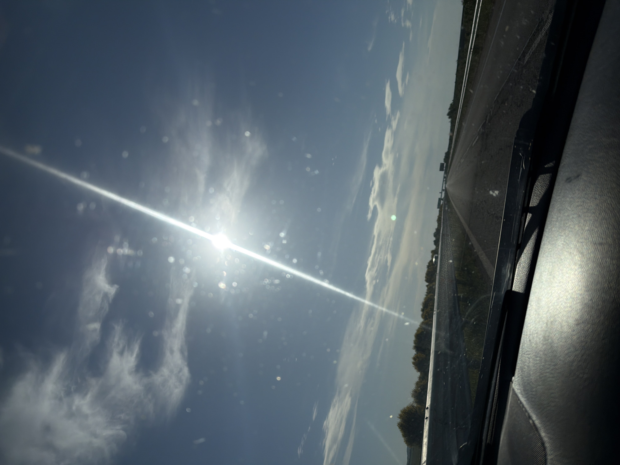The image shows a view from inside a car, looking out onto a highway. The sun is bright in the sky, with visible rays and some scattered clouds. Part of the car's dashboard and windshield wiper are visible in the foreground. The highway stretches into the distance, flanked by trees and vegetation. The location cannot be determined without additional information.