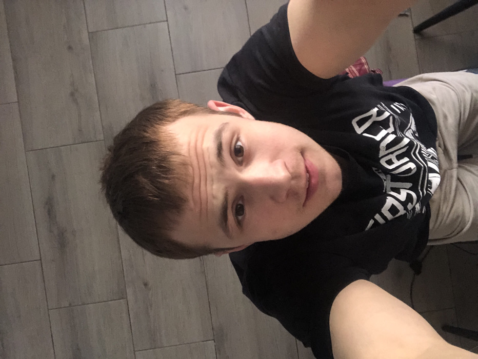 The image captures a selfie of a young person from a high-angle perspective, rotated ninety degrees clockwise. The person, who appears to be male, is looking upwards directly at the camera with a slight, friendly smile. They have short, brown hair, and there are several horizontal lines visible on their forehead, likely due to their expression or the camera angle. Their eyes are brown.
The individual is wearing a black short-sleeved t-shirt with stylized white text and graphics on the front. Parts of the text are visible, including what appears to be "BEST" at the top and a larger, more complex design below it that contains letters suggestive of "DESTROYER" or a similar word, possibly part of a brand or graphic. They are also wearing light grey or beige sweatpants or joggers.
The background primarily consists of a light grey plank-style floor, indicating an indoor setting. The floor planks are laid out horizontally across the current orientation of the image. In the upper right corner, a dark, vertical object, possibly a furniture leg, is partially visible. A small hint of a purple object, which could be a mat or cushion, is also faintly seen behind the person's back. The overall impression is one of a casual, relaxed moment.