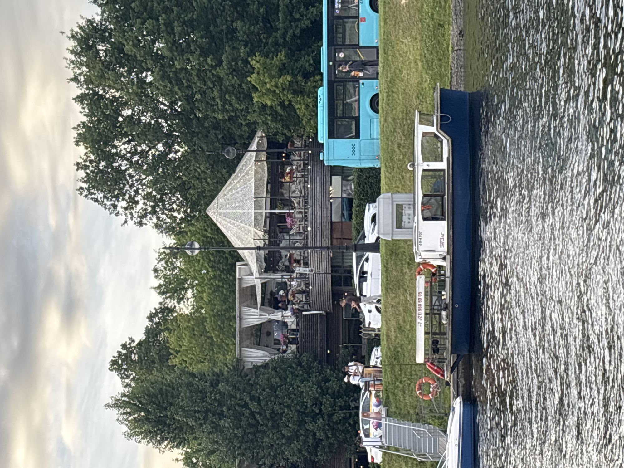 The image captures a waterfront scene in Saint Petersburg, Russia, likely during daylight hours with overcast skies. In the foreground, the dark blue water of a river or canal reflects the sky and surrounding environment. Moored to the embankment is a small, dark blue and white boat. The embankment is lined with lush green grass and a stone retaining wall.
Further back, on the elevated embankment, a blue bus is parked. Next to the bus is a multi-story building that appears to be a restaurant or cafe. It has a rustic wooden exterior and a covered outdoor seating area with white drapes. People are visible sitting at tables on this upper level, suggesting a leisurely atmosphere. Below this restaurant, a few white cars are parked on a paved area, and a set of metal stairs leads down to the embankment. A lamppost stands near the restaurant.
On the embankment, near the stairs and cars, several people are gathered. One individual is walking away from the cars, carrying something. Two other individuals are standing near a small white structure on the embankment. Another two figures are standing near the blue bus. On the moored boat, there are visible life preservers. The overall impression is a lively, public area along a waterway in an urban setting.