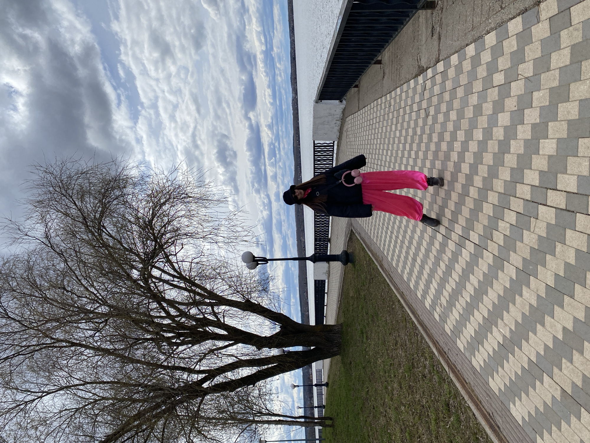A woman stands on a paved walkway beside a body of water in Votkinsk, Russia. She is wearing a black jacket, bright pink pants, a black hat, and carrying pink headphones. A large, bare tree is to her left. Street lamps and a black railing line the walkway, separating it from a grassy area that slopes down to the water. The sky is cloudy with patches of blue. The walkway is paved with alternating light and dark grey rectangular stones.