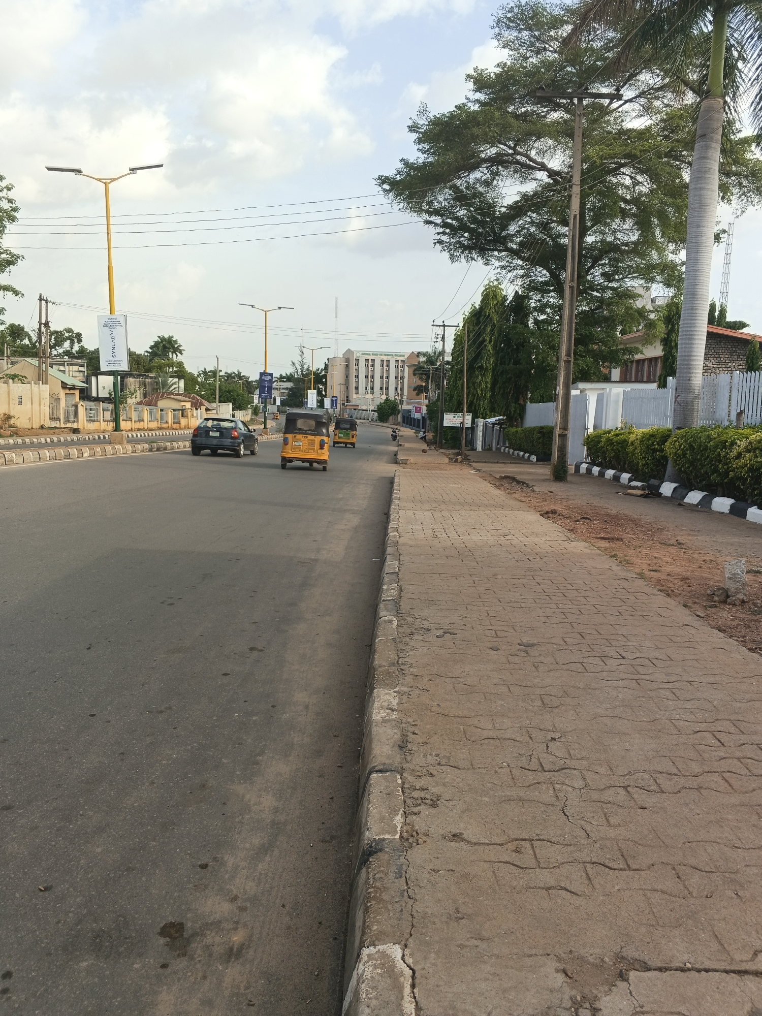 The image depicts a street scene in Shaba, Nigeria. The asphalt road curves slightly to the left and features a black car and two yellow auto rickshaws (tricycles) moving along it. The sidewalk is made of interlocking concrete pavers and runs along the right side of the road, bordered by low shrubbery and a white fence. Tall trees with green foliage line the sidewalk. Overhead power lines are visible. Streetlights with yellow poles are placed along the road. A multi-story building is visible in the background.