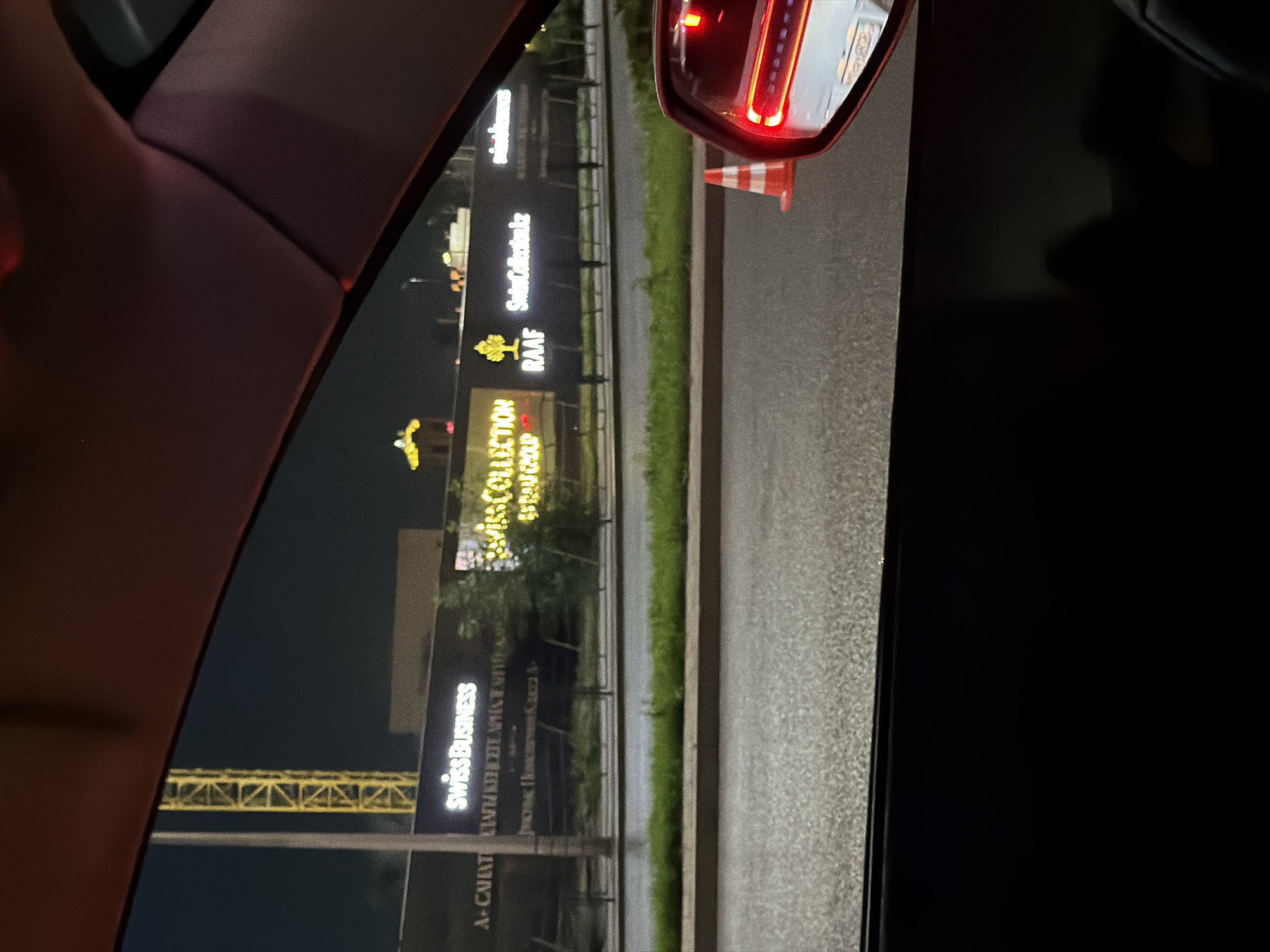 The image, taken at night in Astana, Kazakhstan, shows a view from inside a vehicle looking outwards. A large billboard is visible with illuminated text, displaying "SwissBusiness" and "SwissCollection." Below these, there are other words in Cyrillic, possibly indicating an address or company information, and the website "SwissCollection.kz." To the left, a yellow construction crane is silhouetted against the dark sky. In the foreground, a traffic cone is visible next to a car's side mirror, which reflects the taillights of another vehicle. The road surface is wet, suggesting recent rain. Green grass borders the road in front of the billboard.