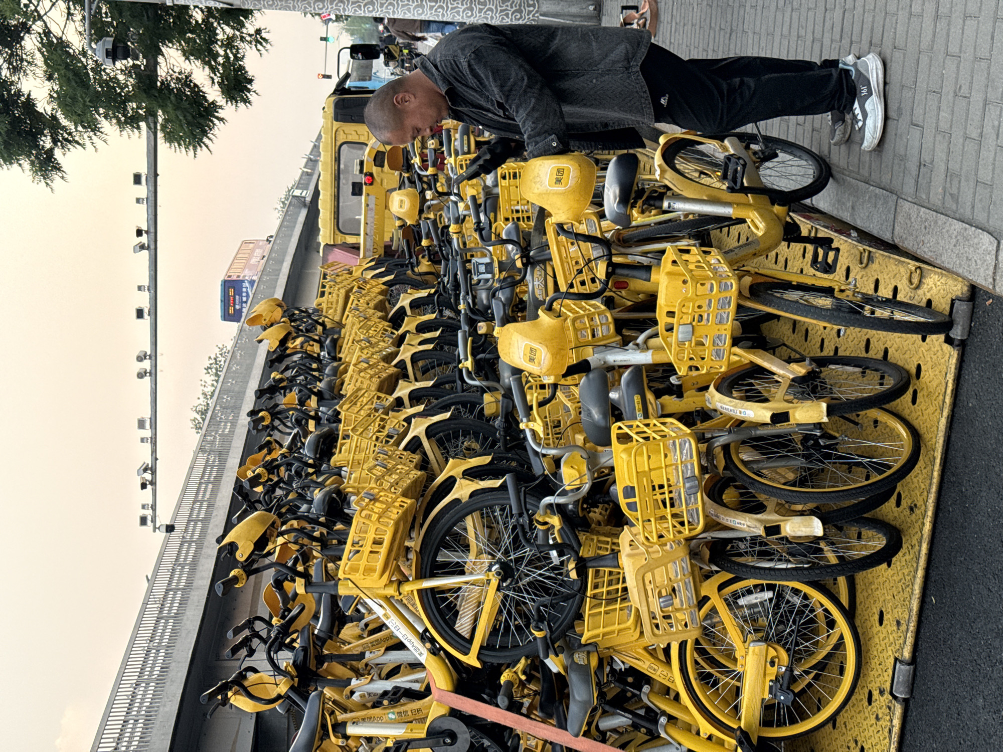 A man is working on a truck filled with yellow Meituan bicycles in Dongcheng District, China. The man is bald and wearing a dark gray jacket and black pants. He is leaning over the bicycles, appearing to be inspecting or repairing them. The truck is full of the yellow bicycles, with their distinctive front baskets. In the background, a bridge is visible, along with trees and a gray sky. The sidewalk is to the right of the truck.