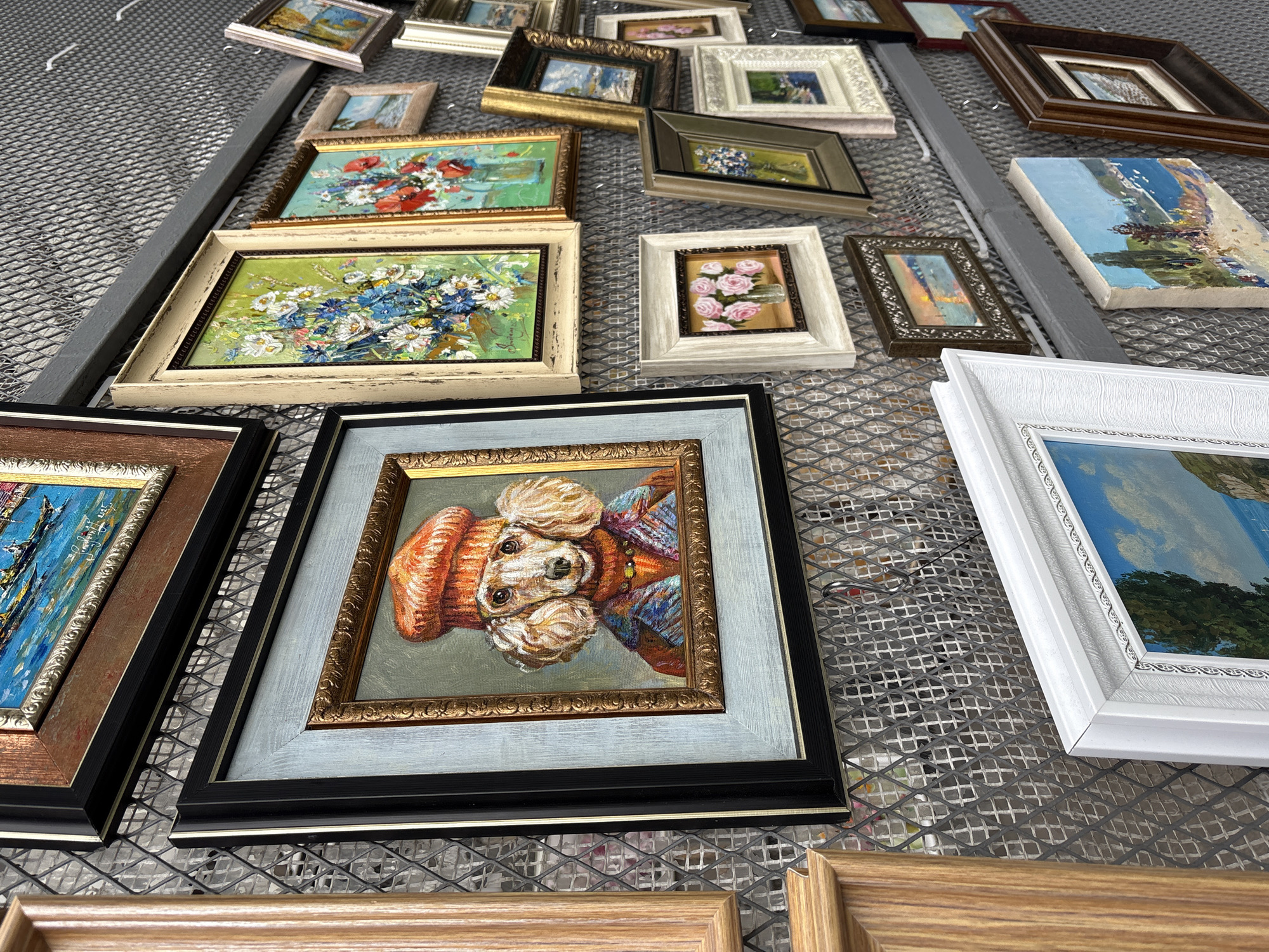 The image shows an assortment of framed paintings laid out on a gray metal grid surface. The paintings vary in size, style, and subject matter, including landscapes, floral arrangements, and a portrait of a dog. The frames are of different materials and colors, including white, black, gold, and wood tones. The setting appears to be an outdoor display, possibly an art market. The location in the city of Moscow, Russia cannot be determined from the image alone.