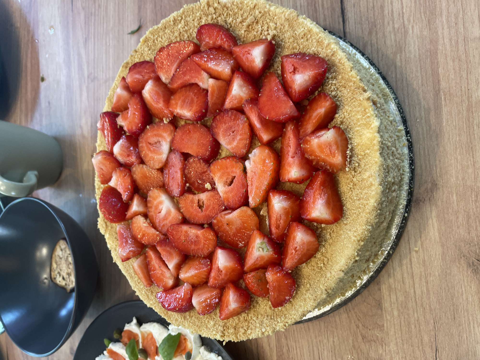 A close-up, eye-level, indoor shot shows a round cake topped with sliced strawberries. The cake is on a dark plate with a metallic rim, which sits on a wooden surface. To the left of the cake, there is a black plate with what appears to be a piece of bread and a black bowl. In the background, out of focus, are two cups. The lighting is natural, coming from the right. The scene is unpopulated and appears to be a domestic setting.