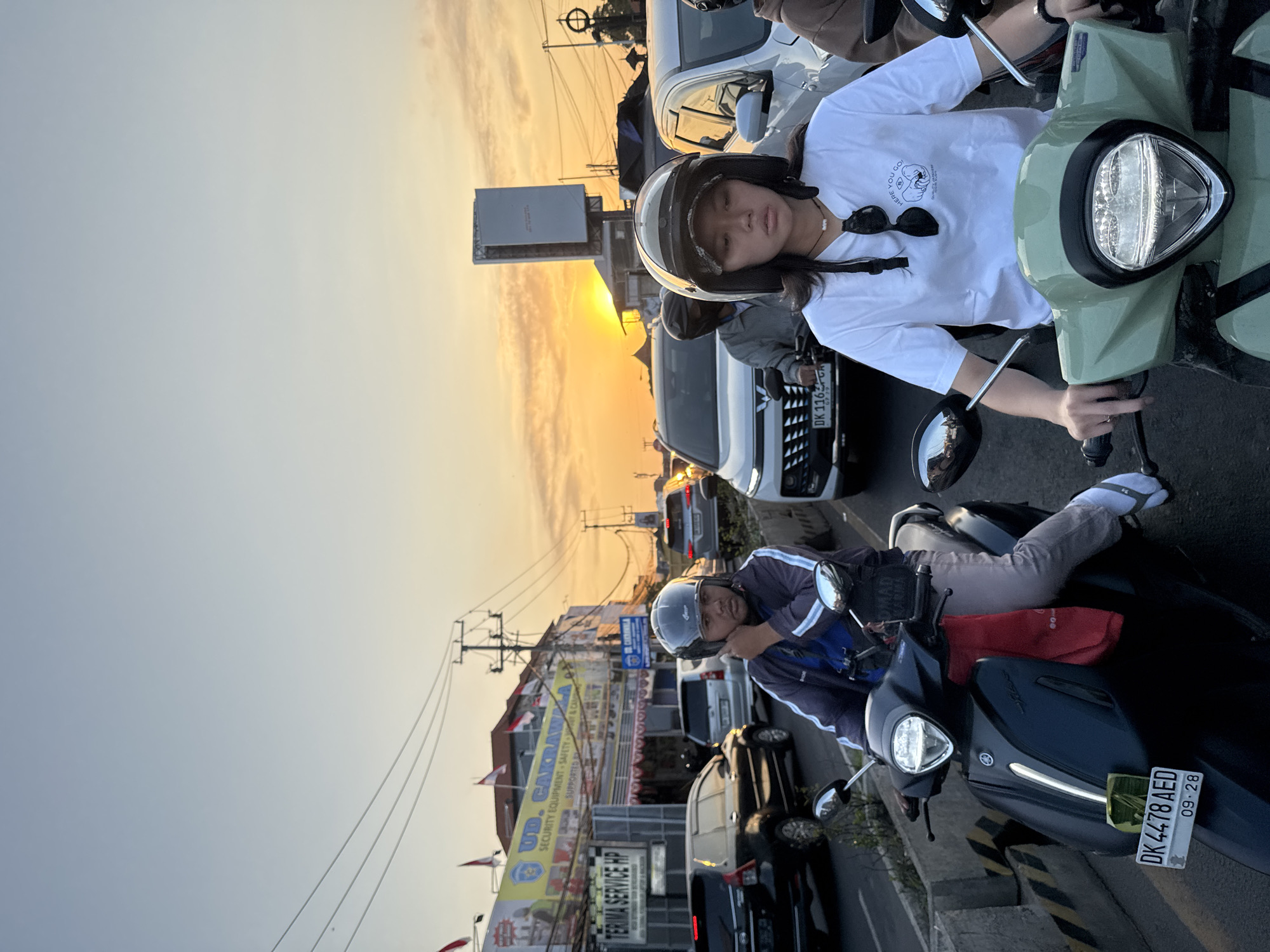 The image captures a street scene in Denpasar, Indonesia, during what appears to be rush hour at sunset. The sky is a gradient of pale blue to warm orange, indicating the sun is low on the horizon. The foreground features two people on motorcycles, positioned side-by-side in traffic.
The person on the left, wearing a dark jacket and a grey helmet, is looking downwards and appears to be speaking on a phone. Their motorcycle is dark blue with a red cloth draped over it, and a license plate reading "DK 4478 AED" and "09.28."
The person on the right, a young woman wearing a white t-shirt and a patterned cap under her helmet, is looking forward. She has sunglasses hanging from her t-shirt and a necklace. Her motorcycle is light green.
In the background, a line of cars and other motorcycles are visible, indicating a traffic jam. Buildings line the street, one of which has a large yellow sign with Indonesian text and logos. Power lines and utility poles are prominent overhead. The overall atmosphere is that of a busy urban environment transitioning from day to evening.