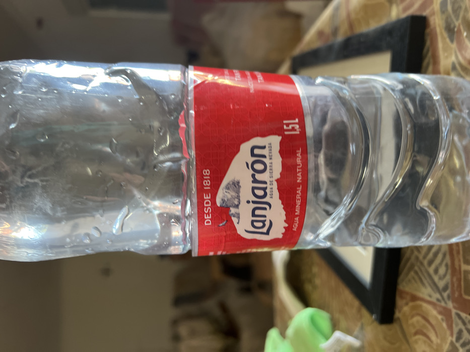 The scene depicts an indoor setting in La Coma, Spain. A transparent plastic bottle, approximately half full with clear liquid, stands prominently in the foreground. The bottle's exterior is covered with small water droplets, indicative of condensation. A red label wraps around the middle of the bottle. The label features white text, with "DESDE 1818" at the top, followed by the brand name "Lanjaron" in blue stylized script, and "AGUA DE SIERRA NEVADA" underneath. Below this, "AGUA MINERAL NATURAL" is printed, and "1,5 L" indicates the volume to the right. A small illustration of a snow-capped mountain is visible on the label.

The bottle rests on a surface, likely a table, covered by a patterned textile in shades of brown, beige, and possibly dark red. To the left of the bottle, a partial view of an irregular, neon green object is present. To the right, a dark, rectangular object, consistent with a picture frame, is partially visible. The background is significantly out of focus, revealing indistinct shapes suggesting blurred furniture elements, such as chair backs or armrests, and possibly walls. No individuals are visible, and no actions or interactions are occurring.