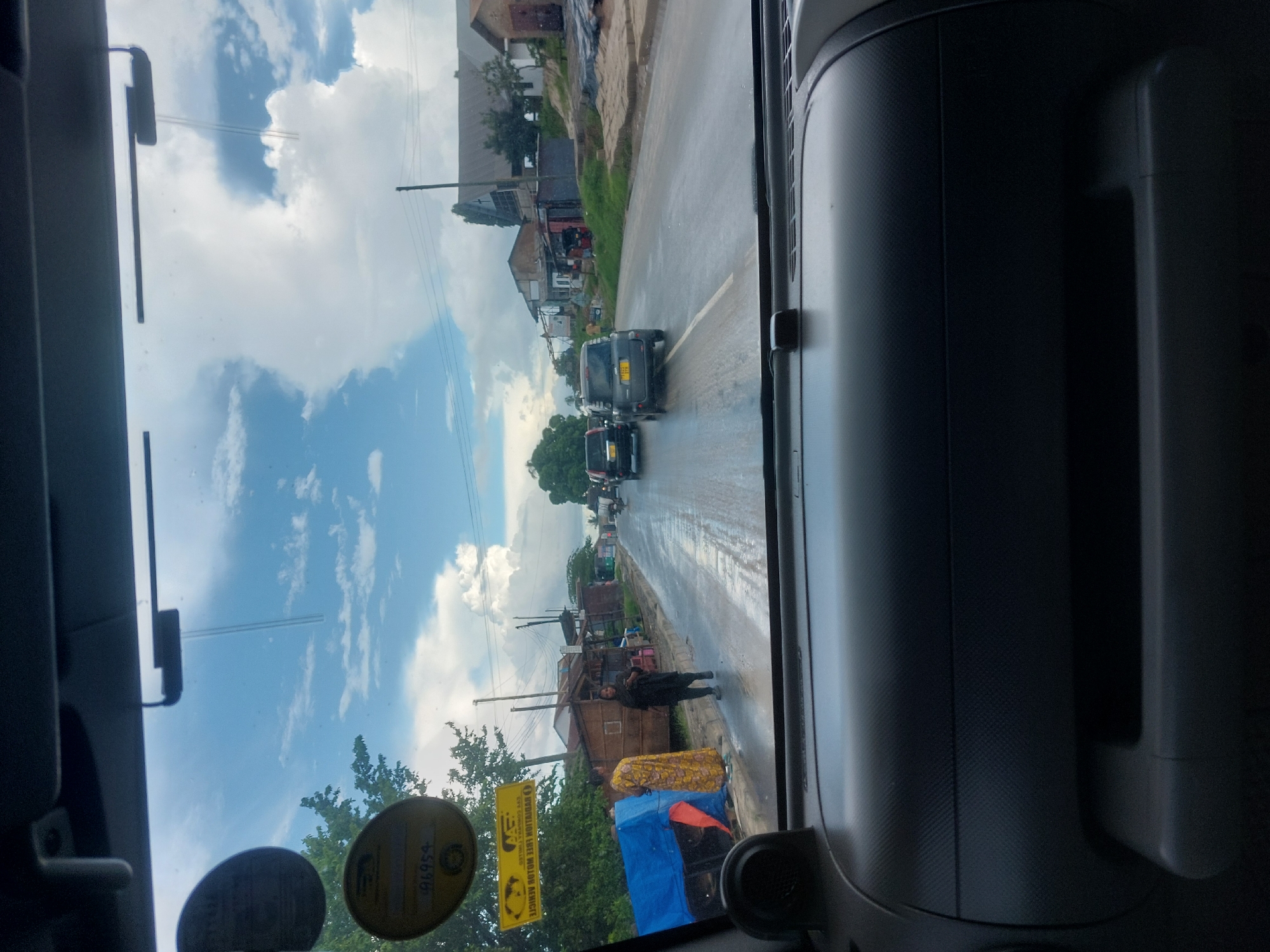 The image, taken from inside a vehicle, shows a street scene in Dar es-Salaam, Tanzania. A line of traffic is heading up the road. On the right side of the road, a person is standing beside blue tarpaulin-covered merchandise, possibly for sale. The weather is overcast, with clouds in the sky. Buildings line the street. Yellow advertising is placed on the left side of the image.