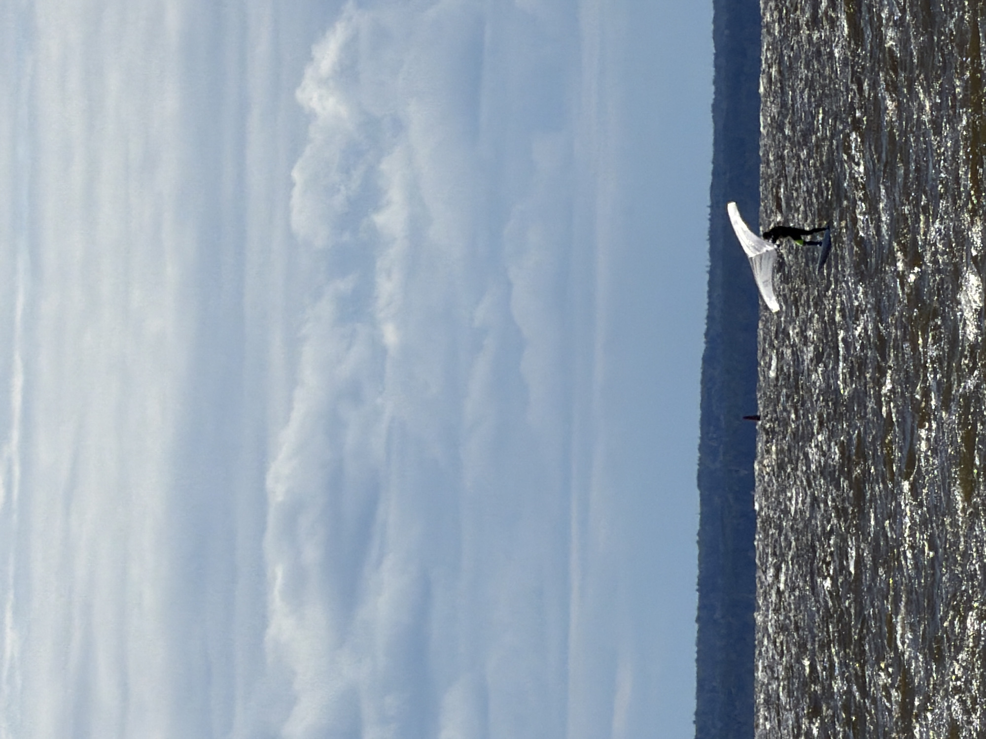 A lone figure is wing foiling on a choppy, dark body of water under a cloudy sky. The individual, wearing a black suit with neon green accents, stands on a board and holds a white wing above their head. The water is illuminated by sunlight, creating a shimmering effect. In the background, a dark, treed shoreline is visible, with a small red buoy further out in the water. The sky is filled with layers of light and dark clouds. The scene is taking place on the Gulf of Finland near Kronstadt, Russia.