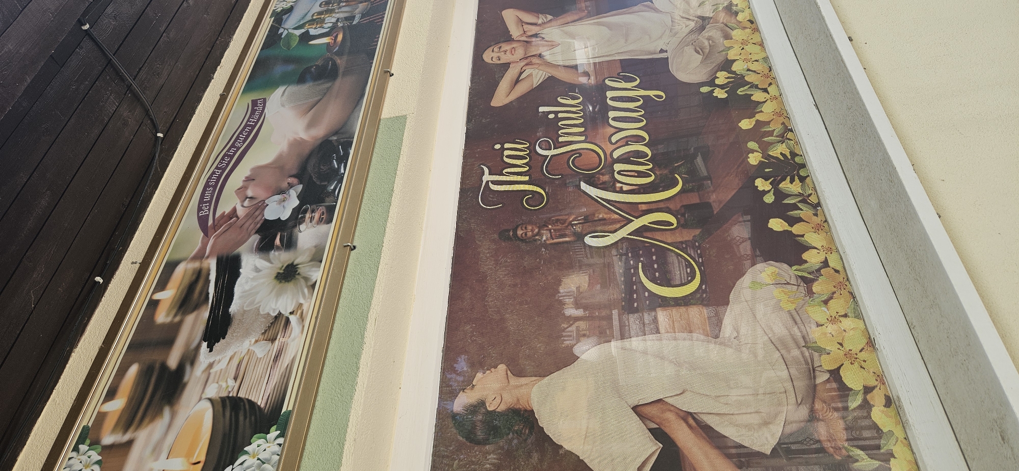 Two framed advertisements are mounted on a wall. The upper advertisement features a woman lying down, with her eyes closed and hands gently massaging her temples. Candles and white flowers are visible in the foreground. A banner above her reads "Bei uns sind Sie in guten Händen," which translates to "With us, you are in good hands." The lower advertisement depicts a woman in profile, facing left, with her eyes closed and her head tilted back. She is wearing a light-colored outfit. To her right, large, stylized text reads "Thai Smile Massage." Behind the text and the woman, a wooden statue of a figure in traditional attire and floral elements are visible. The advertisements appear to be for a massage business. Based on the context provided, this scene is located in Hévíz, Hungary.