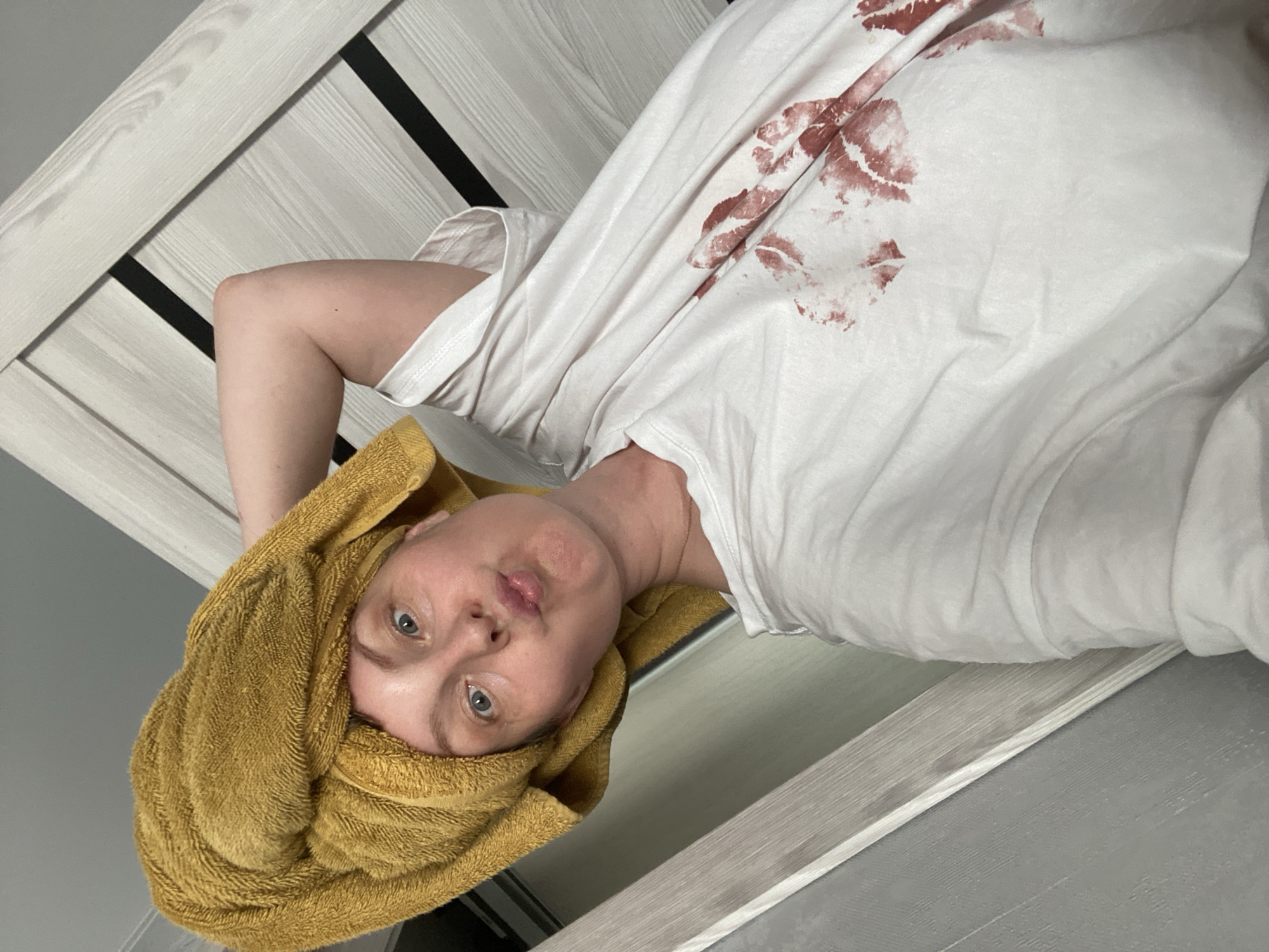 A fair-skinned woman with blue eyes and a yellow towel wrapped around her head poses for a picture. She is wearing a white t-shirt with red lipstick stains on it. The background includes a white and grey patterned door and a grey wall. The location is Noviy, Russia.