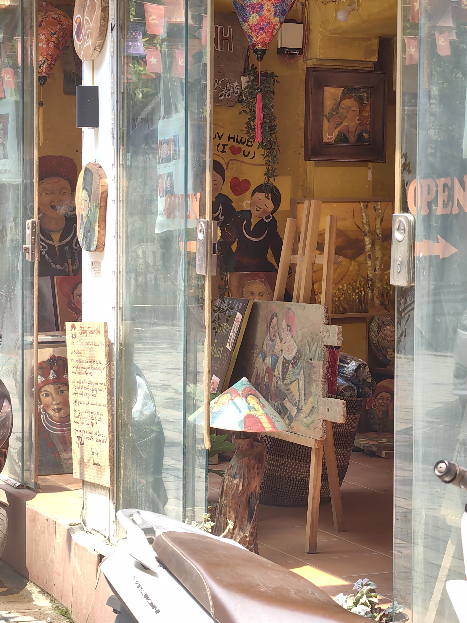 The image depicts the street-level entrance of an art gallery or handicraft shop in Sa Pa, Vietnam, viewed from the exterior. A white scooter with a brown seat, bearing "VIETNAM" on its side, is partially visible in the foreground, parked in front of the establishment.
The shop entrance consists of clear glass doors, through which the interior is visible, with reflections partially obscuring some details. An "OPEN" sign with a right-pointing arrow is affixed to the inside of the rightmost glass door.
The interior walls are painted a uniform yellow. Above the doorway, a string of small red flags with yellow stars, consistent with the Vietnamese flag, is displayed, alongside some red flags featuring a yellow hammer and sickle. Various artworks, primarily paintings and carved wooden pieces depicting people, dominate the display. On the left, a large carved and painted wooden figure of an elderly woman, seemingly smoking a pipe, is visible. Below it, a painting of a young girl wearing a red headscarf is displayed next to a vertical wooden sign titled "Hello From Lan Anh – an artist in Sa Pa!", stating the works are "original artworks – all by hand."
In the center, a wooden easel holds a painting of two figures, possibly women in traditional dress, one with a conical hat. A decorated conical hat rests on a wooden base nearby. Behind the easel, a painting of trees with white trunks and yellow leaves is visible. A large woven basket filled with textiles or other items is positioned further inside. A framed painting of a woman with a head covering smoking a pipe hangs on the yellow back wall. Colorful paper lanterns, one purple and one orange, hang from the ceiling. Green foliage trails down near a lantern. Handwritten text, including "I LOVE SA PA" and hearts, is visible on the upper wall. No individuals are present in the immediate scene.