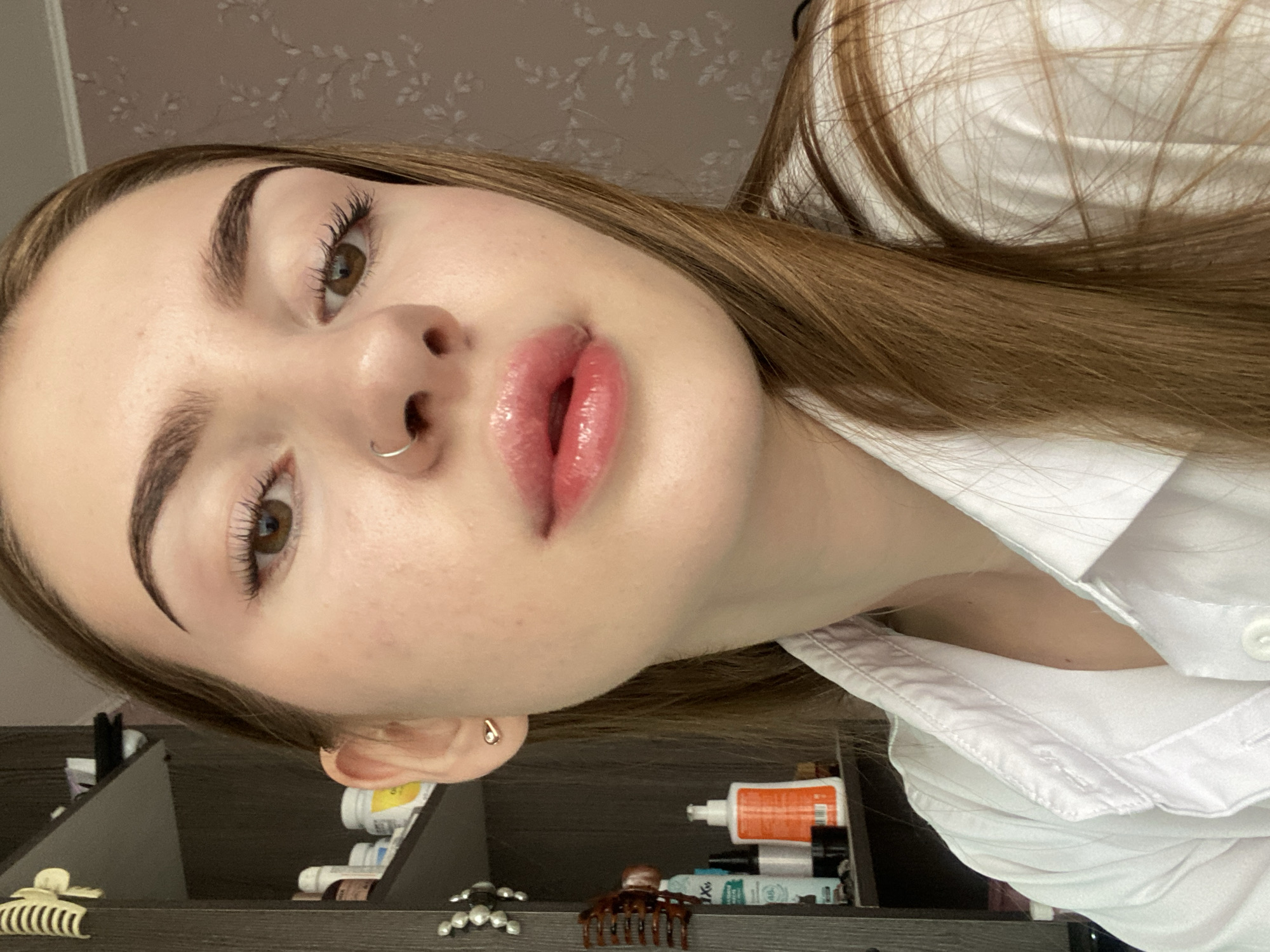 A young woman with long, light brown hair and light skin is shown in a close-up portrait. She is wearing a white collared shirt. She has large, dark brown eyes, dark arched eyebrows, and plump lips with a pink gloss. A silver hoop is visible in her nose, and a small, gold earring with a diamond accent is in her right earlobe. The background is a blurred interior scene, with shelves containing various bottles and items on the left, and a floral patterned wallpaper on the right. No specific location in Dyakovo, Russia is discernible from the image.