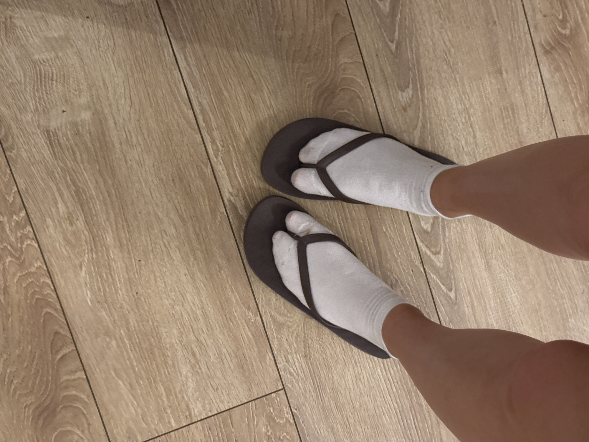 The image shows a pair of feet wearing white socks and dark brown flip-flops, standing on a light brown wooden floor. The floorboards are arranged horizontally in the image and show a wood grain pattern. The feet are positioned slightly apart, with the toes visible through the flip-flop straps. The lighting suggests an indoor setting. The location in Aksu, Turkey is not discernible from the image.