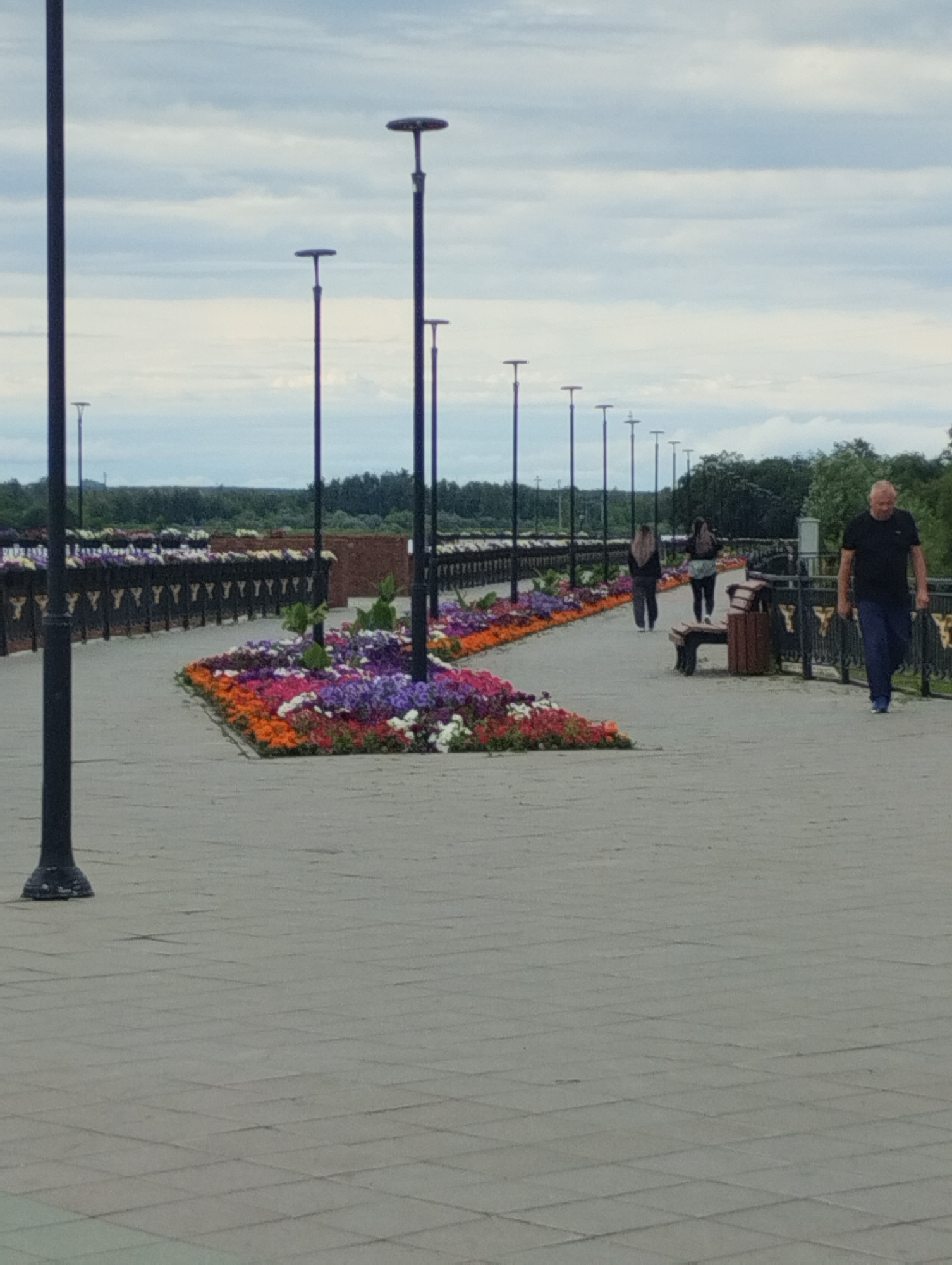 The image depicts a paved walkway with a curving flowerbed in the foreground. The flowerbed is filled with a variety of colorful flowers, including red, orange, purple, pink, and white varieties. Several tall, dark lampposts line the walkway. In the background, a row of dark planters filled with more flowers stretches into the distance. Beyond the planters, a line of trees forms a dark green horizon against a cloudy, pale blue sky.
On the walkway, three individuals are visible. A man wearing a black t-shirt and blue jeans walks towards the right side of the frame. Two women, one in a black headscarf and dark clothing, the other in lighter clothing and a backpack, walk away from the camera towards the left. A park bench and a trash receptacle are situated on the walkway near the women.
The scene is likely a public park or promenade in the city of Kostanay, Kazakhstan. The style of the lampposts and the general landscaping suggest a well-maintained urban green space. The weather appears overcast, but there is no visible precipitation.