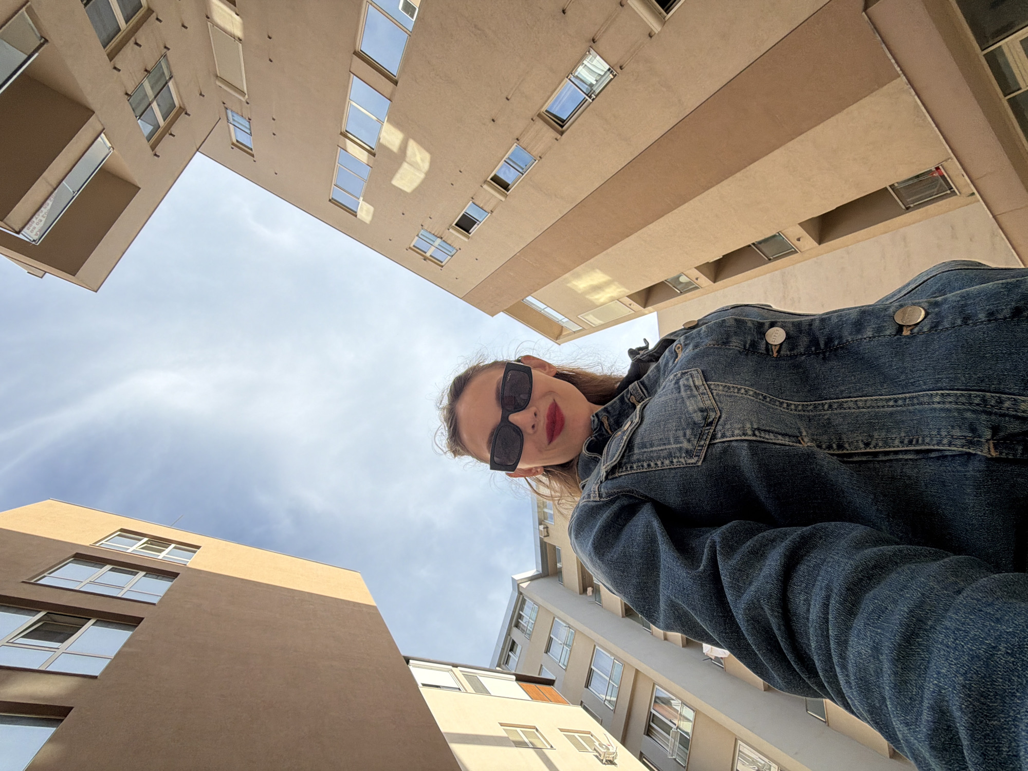 An upward-facing, low-angle shot captures a young woman and surrounding multi-story buildings against a bright, partly cloudy sky, characteristic of an urban setting like Budva, Montenegro. The woman, positioned in the lower right, looks directly at the camera with a slight smile. She wears dark rectangular sunglasses, red lipstick, and a blue denim jacket with "LEVIS" visible on its metal buttons. Her long brown hair frames her face.
The buildings feature light brown/beige facades and numerous rectangular windows, some of which appear open. Two prominent buildings frame the upper and lower left of the image, appearing to lean inwards due to the wide-angle, low perspective. The sky is a clear light blue with scattered wispy clouds, indicating bright daylight, likely late morning or afternoon, and good weather. The composition emphasizes the towering verticality of the surrounding structures.