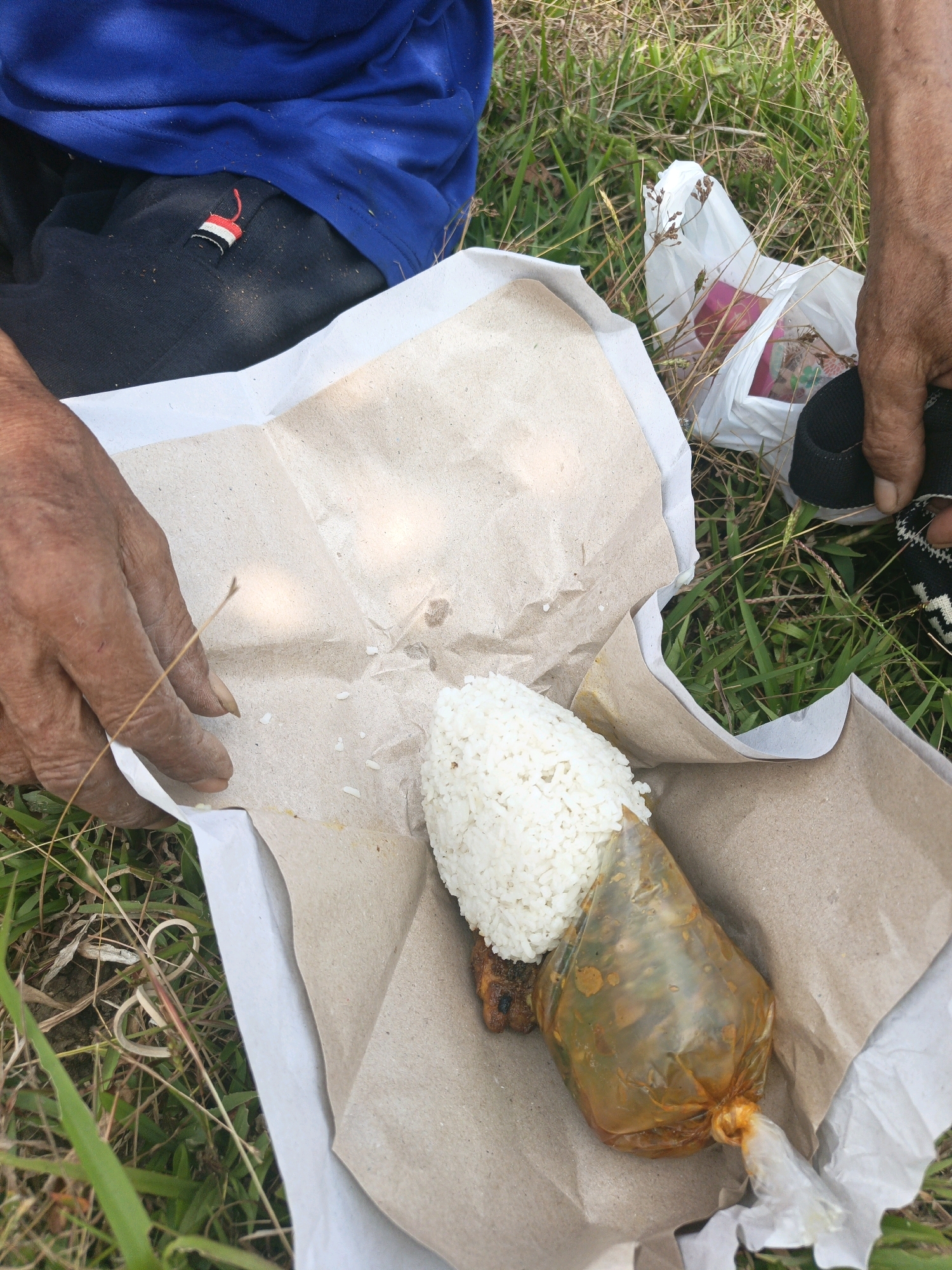 In Sidodadi, Indonesia, a person sits outdoors on the grass. They are wearing a blue t-shirt and dark pants with a red, white, and black striped detail on the left side. In front of them, a meal is laid out on brown paper. The meal consists of a mound of white rice and a piece of meat, both wrapped in clear plastic. The plastic packaging is tied at the bottom. A white plastic bag with items inside, possibly food wrappers, is visible to the right. The ground is covered in dry grass and some green vegetation. The person's hands are visible, one touching the paper wrapping and the other holding the edge of their pants. The scene suggests a casual outdoor meal.