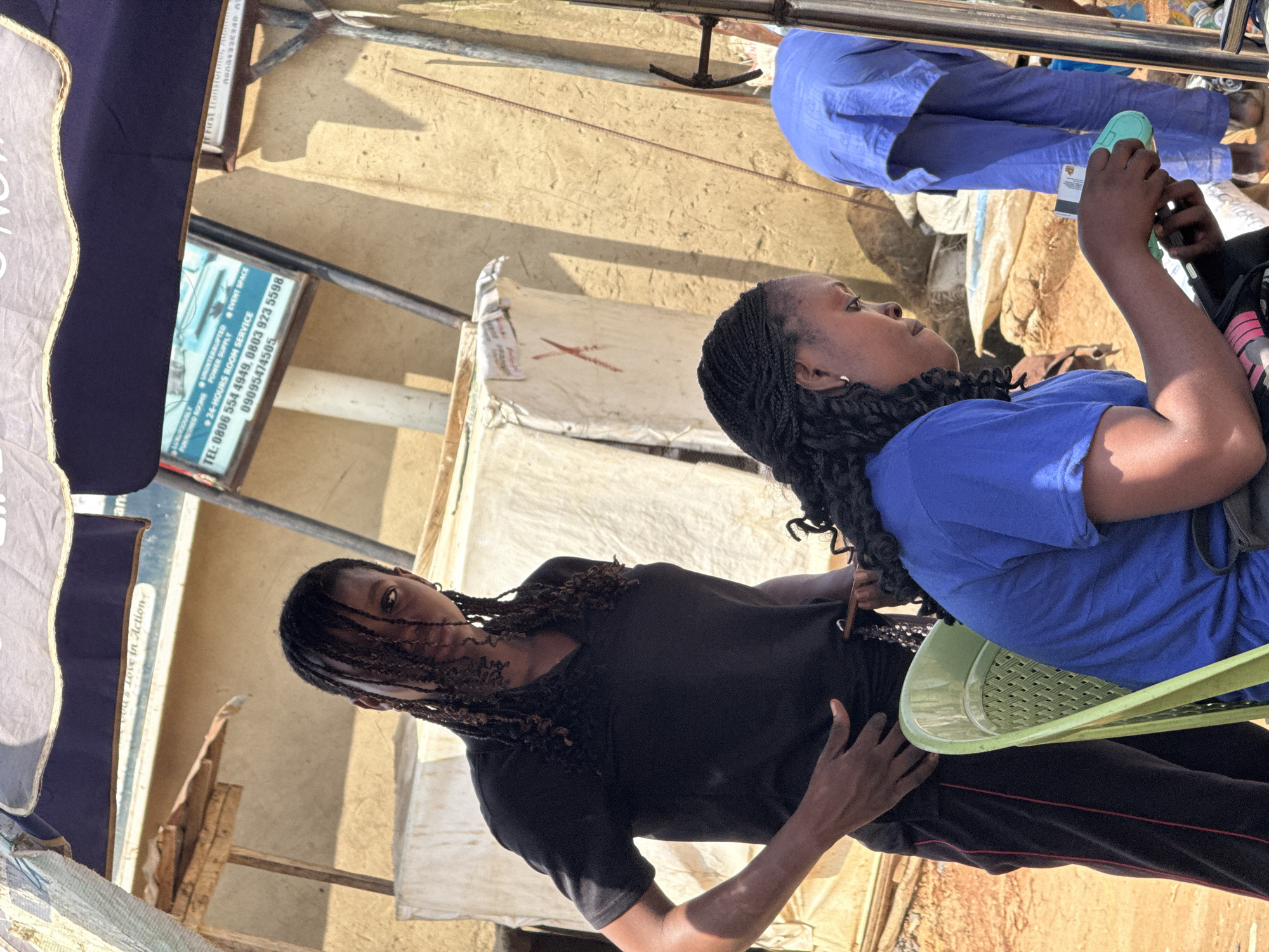 In Abuja, Nigeria, a young woman with braided hair, wearing a black t-shirt and black pants with red stripes, stands and looks towards the camera with her left hand on her hip. To her right, another woman, also with braided hair and wearing a blue t-shirt, is seated and looking down at a cell phone and a mint green object in her hands. A third person, wearing blue pants and a blue top, stands in the background to the right, facing away from the camera. The scene appears to be outdoors, possibly in a market or street setting, with a textured, light-colored wall in the background and a blue awning overhead. Debris and various objects are visible in the surroundings.