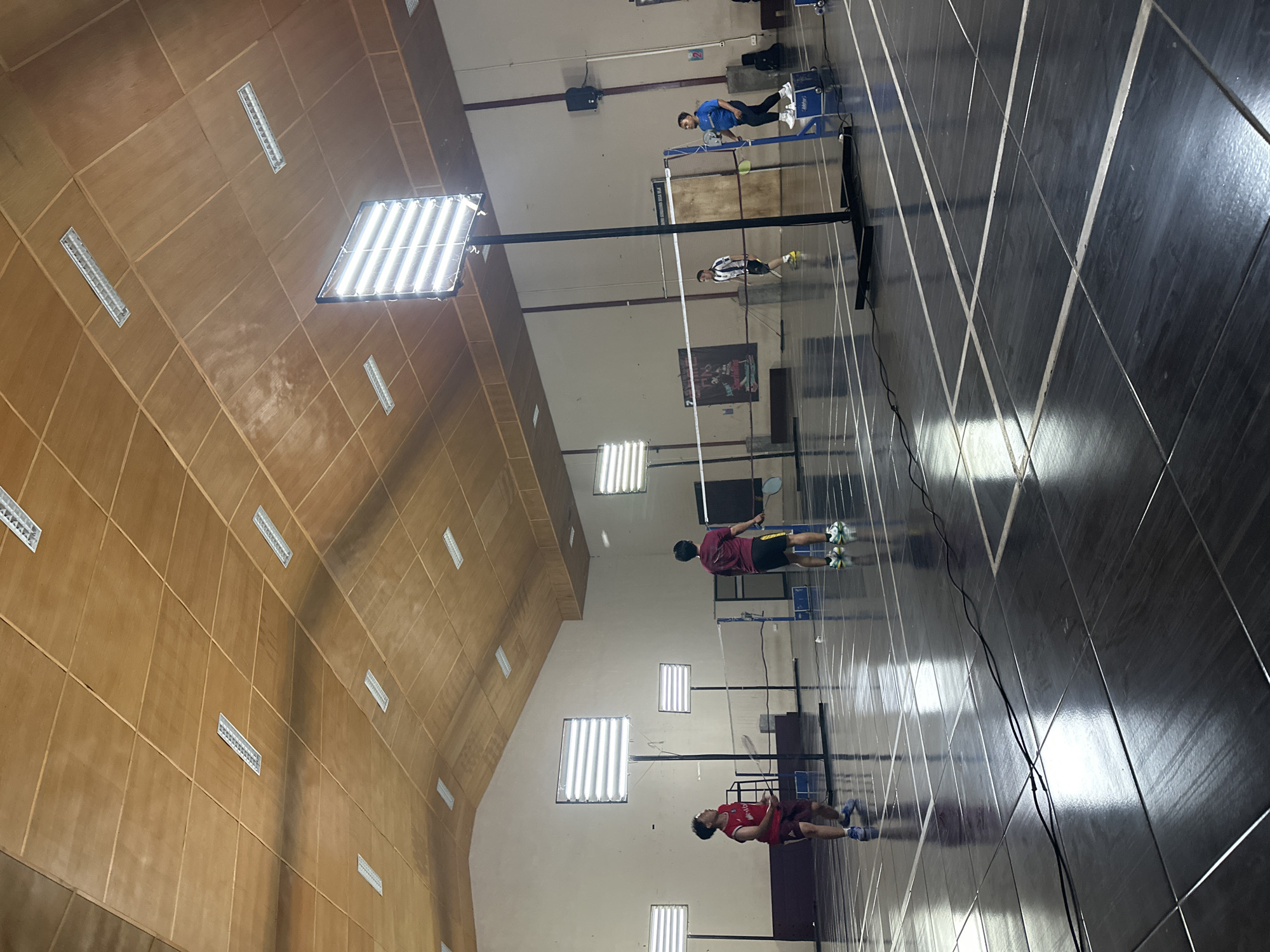 The image depicts an indoor badminton court at GOR Bimasakti in Batu, Indonesia. Four individuals are engaged in a badminton match. In the foreground, two players face each other across the net. One player, wearing a red shirt and dark shorts, is in motion, holding a badminton racket. The other player, in a maroon shirt and black shorts, stands ready to receive. Further back, two more players are visible on the other side of the court. The court itself is well-lit by large, rectangular artificial lights mounted on poles and also by linear fluorescent lights integrated into the wooden paneled ceiling. The polished floor reflects the overhead lighting and the court markings. A black power cord snakes across the floor in the foreground. The environment suggests an active sporting event or practice session.