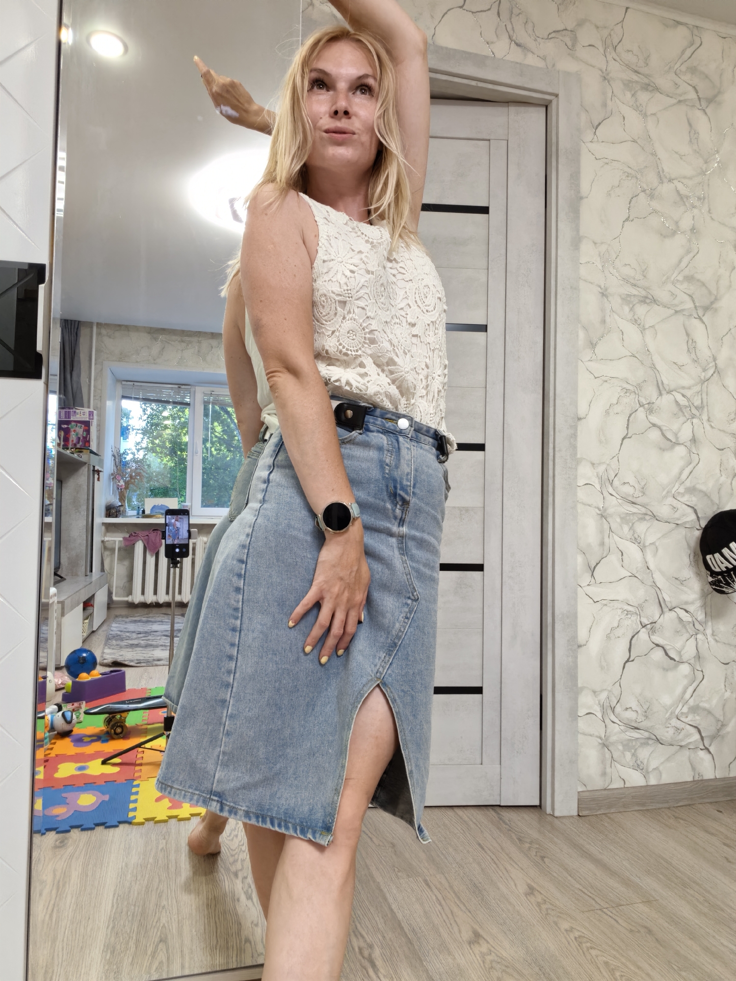 A Caucasian woman with blonde hair is standing in a room, viewed from the chest down and reflected in a large mirror. She is wearing a white lace top and a light blue denim skirt with a slit up the side. Her right arm is raised above her head, and her left hand rests on her hip. A smartwatch is visible on her left wrist. In the mirror's reflection, a phone on a tripod is visible in the background, suggesting she might be recording or taking a selfie. The room appears to be a living space, with a door, patterned wallpaper, and a floor with a wood-like finish. A child's play mat with colorful shapes is visible on the floor in the reflection, along with some toys. The overall impression is of a casual indoor setting. The location is a residential apartment in the city of Sterlitamak, Russia.