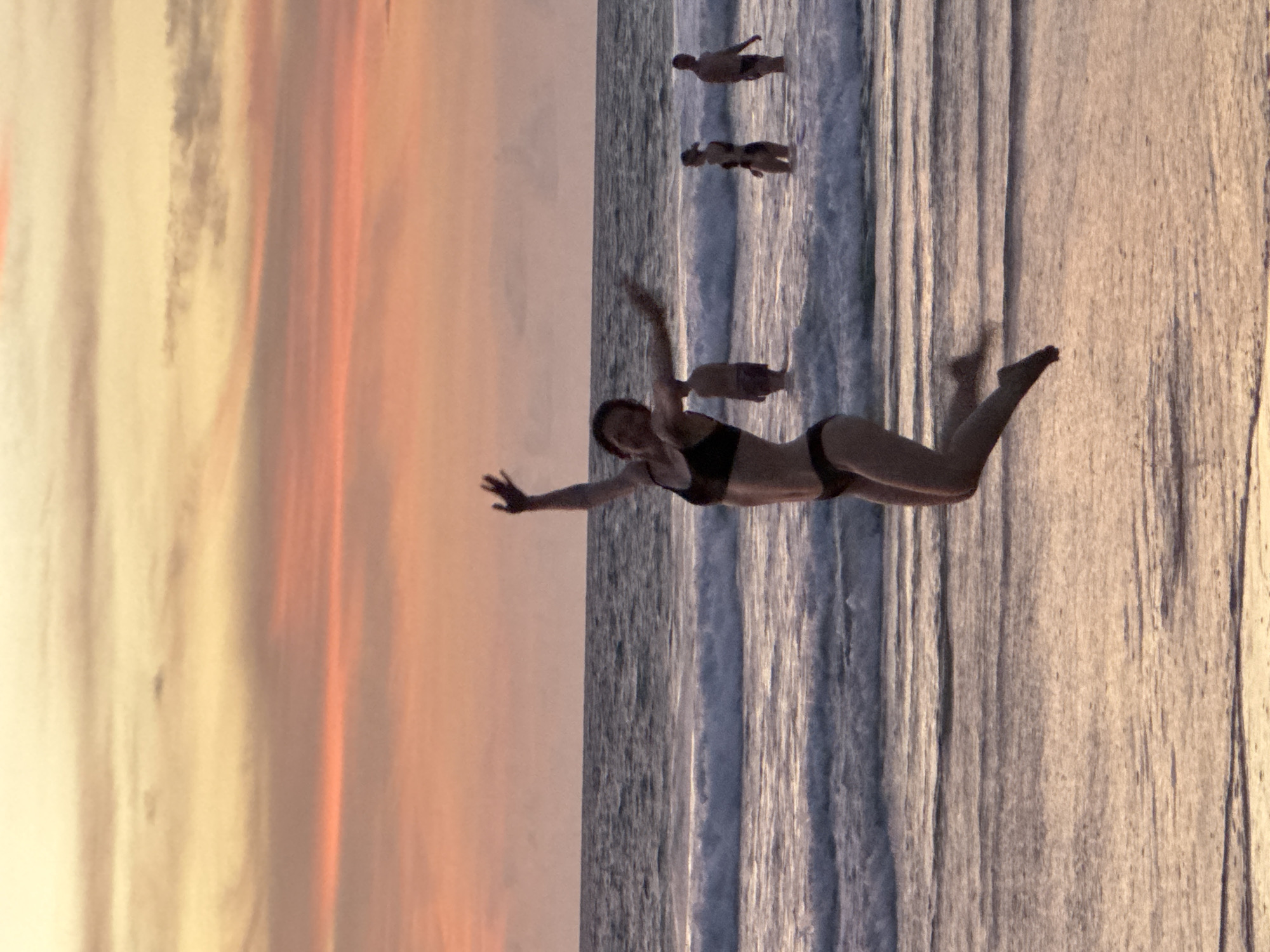 A person is captured mid-air, jumping joyfully on a beach in Karon, Thailand, during sunset. The sky displays a gradient of warm colors, transitioning from soft yellow to pink and orange hues, with horizontal cloud formations. The person in the foreground is wearing a dark bikini and appears to be celebrating, with arms outstretched and legs bent. In the background, several other individuals are visible in the shallow water, standing or wading. Gentle waves are rolling onto the sandy shore. The overall scene evokes a sense of freedom and happiness associated with a beach vacation at dusk.