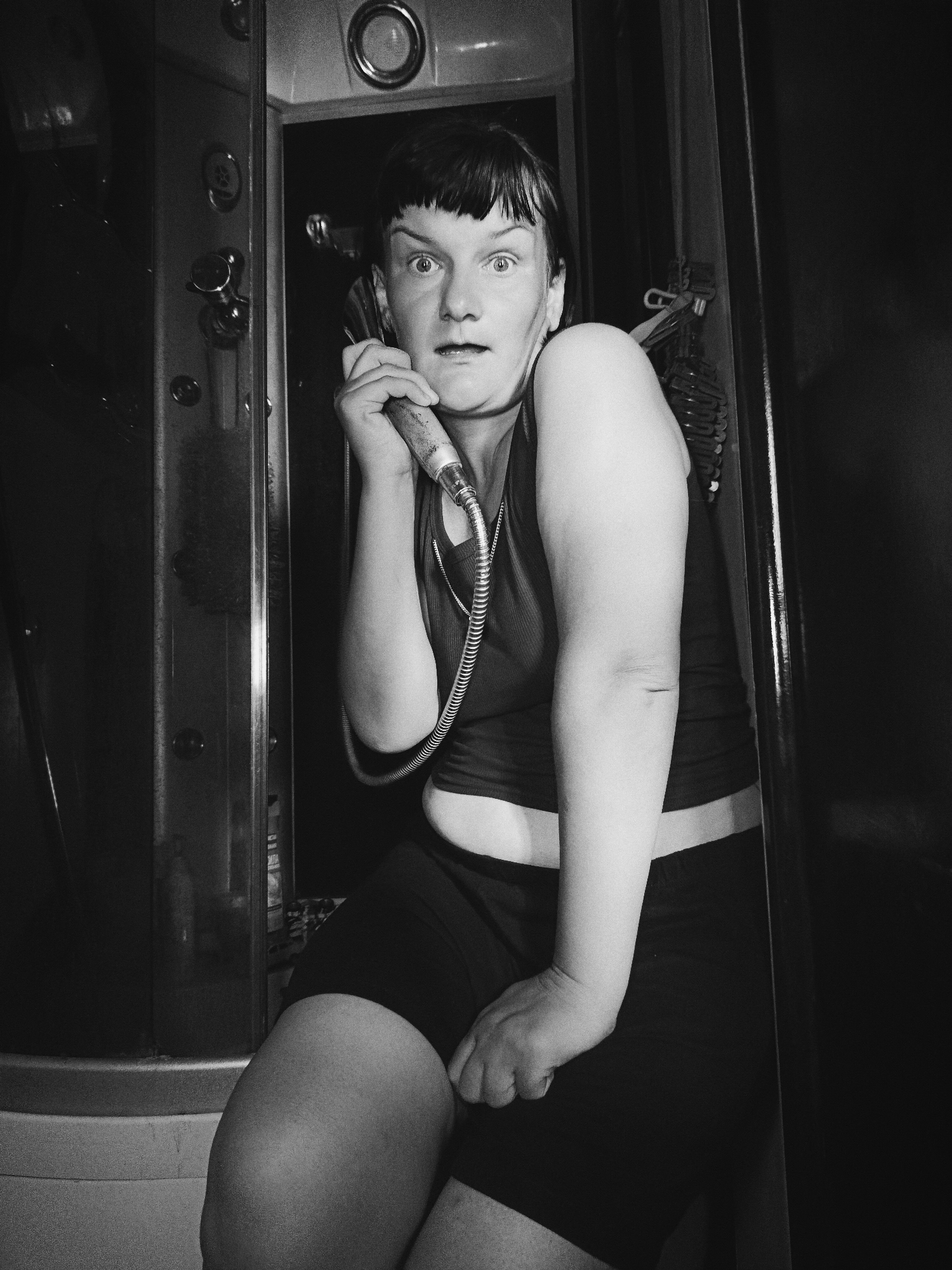 A black and white image shows a person with short, dark hair and bangs, appearing to be seated and holding a shower head to their ear as if it were a telephone receiver. The person is looking forward with a surprised expression. They are wearing a tank top and dark shorts. They appear to be inside a shower cabin, which is partially visible to the left of the person. Hooks and possibly clothing hangers are visible behind the person to the right. The location in Фоки, Russia is not evident from the image.