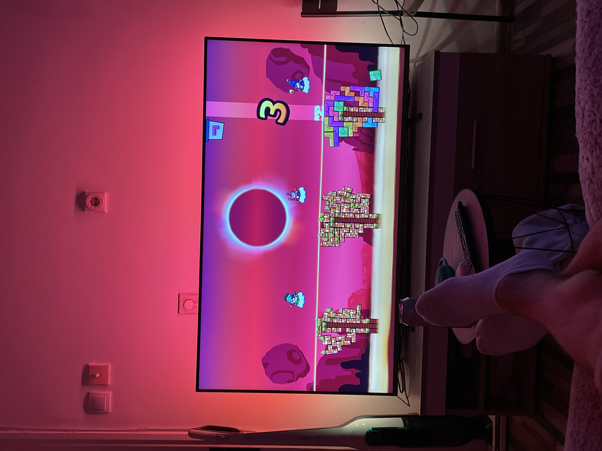 A brightly lit television screen displays a video game featuring a pink and purple landscape with a large glowing circle. The game shows characters flying and structures built from blocks. The screen is on a console in a room with a red-toned wall. Two electrical outlets and a light switch are visible on the wall. A cordless vacuum cleaner rests below the console, and part of a person's leg and foot in a white sock are visible on the right side of the frame. The exact location in Belgrade, Serbia is not discernible.