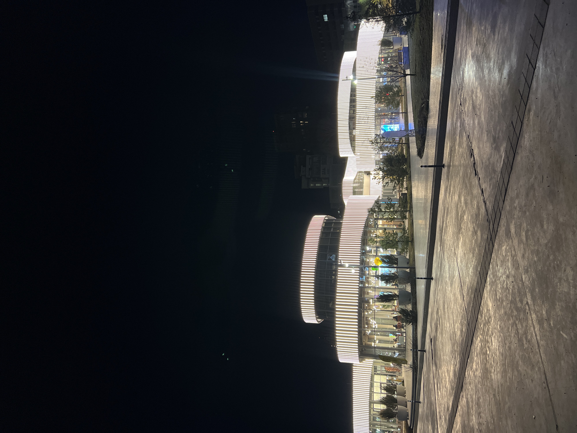 The image shows the illuminated Kazan Mall in Kazan, Russia, at night. The multi-story building features a distinctive circular architectural design with horizontal bands of light. The exterior is lined with windows, suggesting retail or commercial spaces within. A paved walkway extends along the base of the building, with occasional trees and other landscaping elements. The sky is dark, indicating nighttime. There are no visible people.