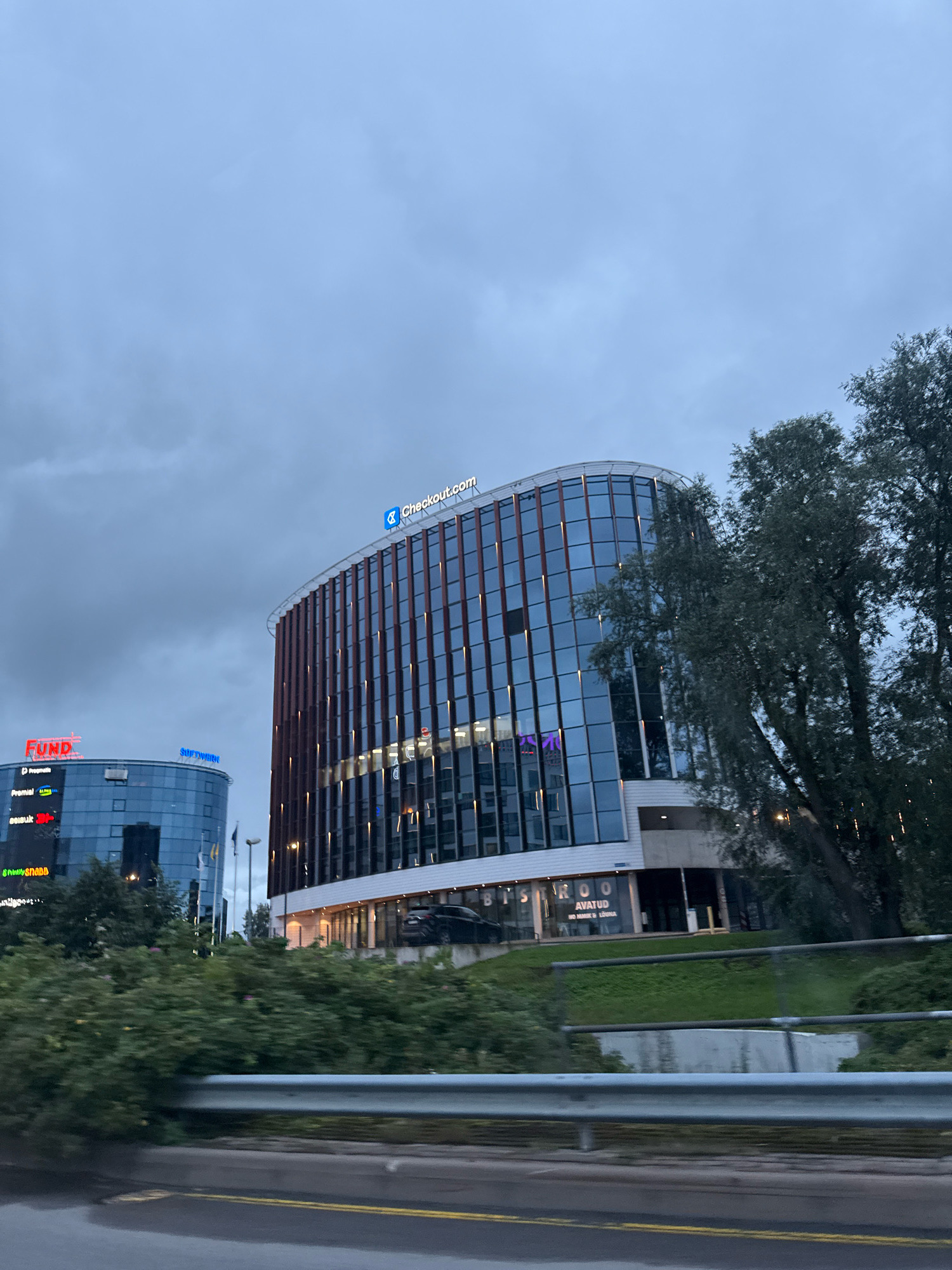 The image shows two modern office buildings in Tallinn, Estonia. The building on the right has a curved glass facade with vertical brown accents and is topped with a "Checkout.com" sign. The building on the left is a darker glass structure with red and blue signage, including "FUND" and "SOFTWERK". The scene is set against a cloudy sky and features trees on the right side of the frame. A road and a metal guardrail are visible in the foreground, suggesting the photo was taken from a vehicle.