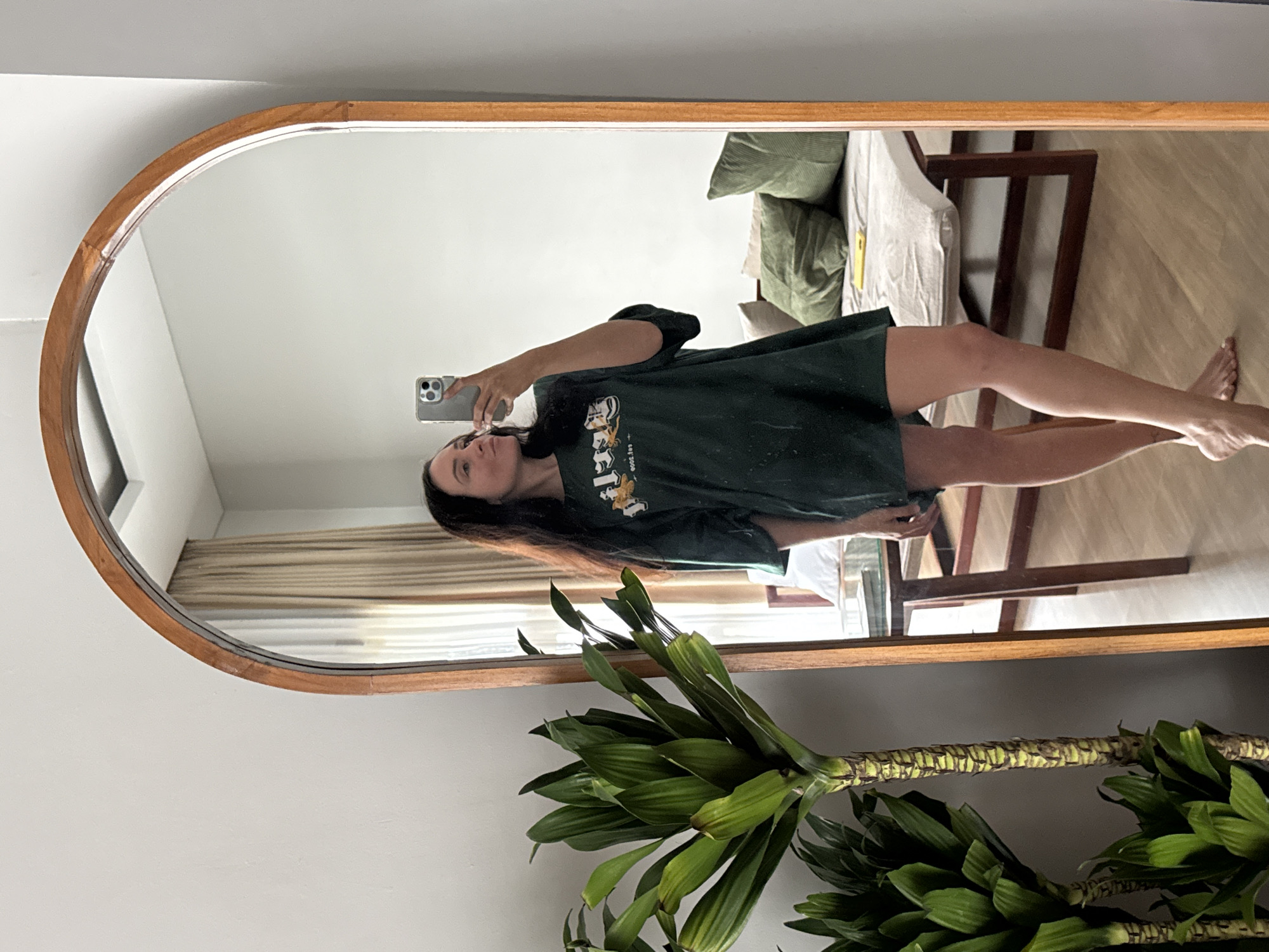 A woman with dark hair is taking a mirror selfie in a room. She is wearing a dark green oversized t-shirt and no pants. Her right hand is holding a silver smartphone, angled to capture her reflection. The background of the mirror shows a bed with green pillows, a table with some items on it, and curtains. To the left of the mirror is a large green potted plant with thick stalks and broad leaves. The floor is light-colored wood. The room appears to be a bedroom or a living area, potentially in North Kuta, Indonesia, given the presence of tropical plants and the general style of the furnishings.