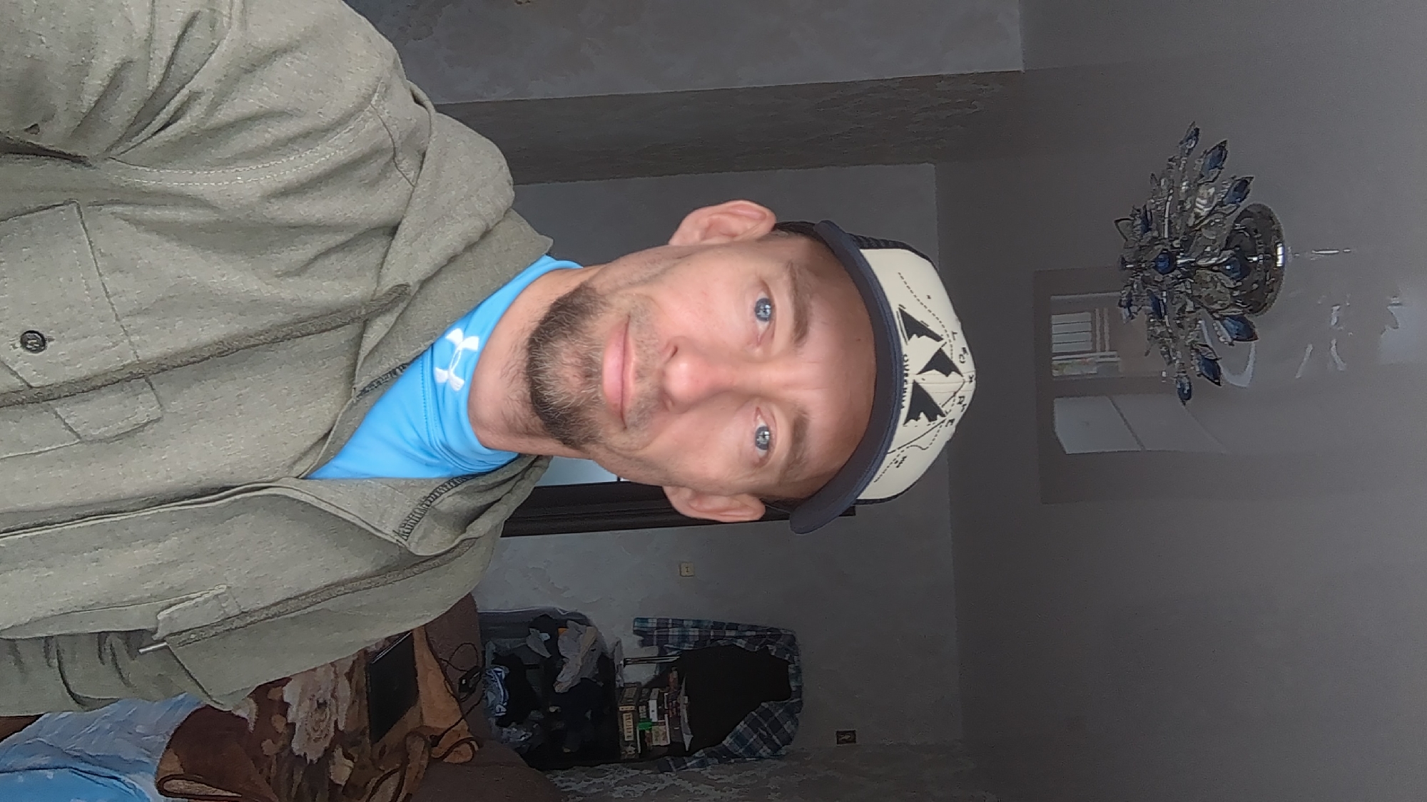 A man wearing a light green jacket over a light blue shirt, and a beige and black baseball cap, is in a room in Ulyanovsk, Russia. A bed with a patterned brown and floral bedspread is visible to the lower left. A pile of items including clothes and boxes sits on the floor in the lower center of the frame. A light fixture with silver and blue elements is attached to the ceiling in the background.