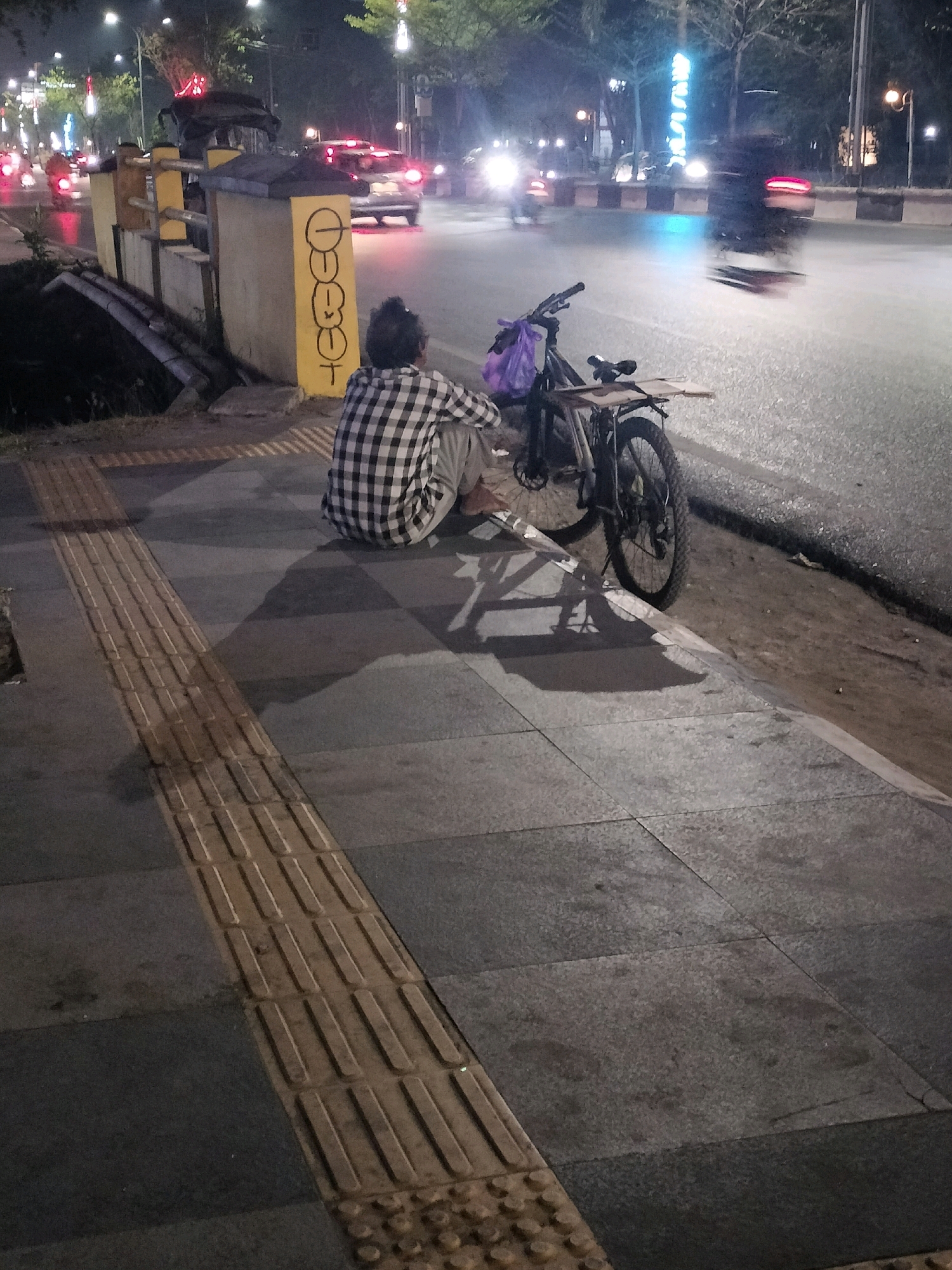 A person wearing a checkered shirt and pants sits on a sidewalk next to a bicycle at night. The person's back is to the camera. The bicycle has a purple bag on the back. The sidewalk is made of gray tiles and has a tactile paving strip. In the background, there is a street with cars and streetlights. There is graffiti on a yellow pillar. The image is taken in Banjarmasin, Indonesia.