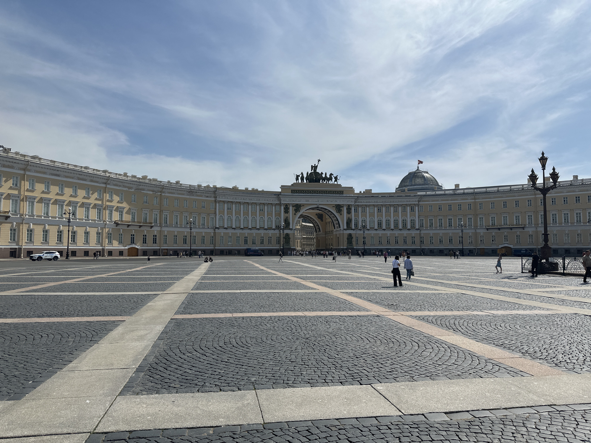 The image captures a wide-angle view of Palace Square in Saint Petersburg, Russia. The square is paved with a pattern of dark, cobblestone-like material interspersed with lighter-colored stone pathways. In the background, a large, curved, light-yellow building dominates the scene. This building features rows of windows and decorative architectural elements. Above the center of the building is a triumphal arch topped with a sculptural group depicting a chariot pulled by horses. A dome-shaped structure with a flag on top can be seen on the right side of the building. Scattered throughout the square are several people, some walking and others standing. On the left side, there is a parked white car. A streetlight is visible on the right side. The sky above is blue with scattered clouds.