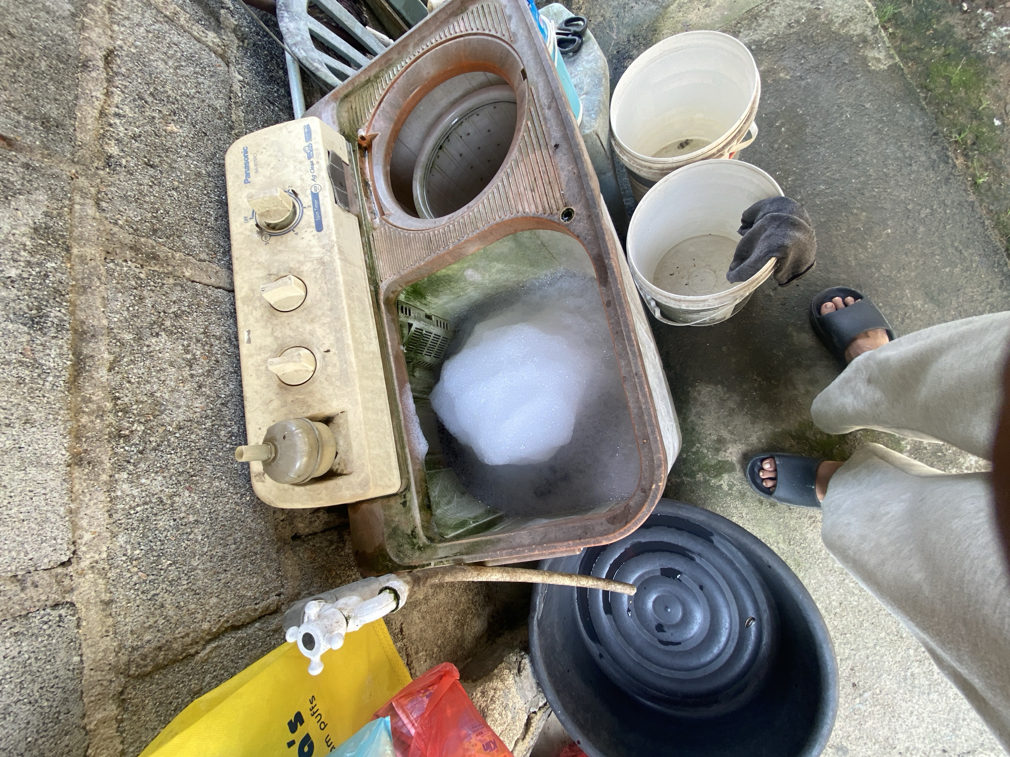 A top-down view captures a weathered, beige Panasonic twin-tub washing machine situated outdoors, possibly in Batam, Indonesia, adjacent to a rough, textured concrete wall. The washing machine features two tubs; the left tub is filled with a significant amount of white suds, indicating recent use. A white faucet with a greenish patina is mounted on the wall above the left tub, with a thin, slightly rusty pipe extending downwards into the tub. Next to the washing machine, to the left, is a large, black plastic basin. To the right of the washing machine are two white buckets, one with a dark grey cloth inside. A person's legs, clad in light grey sweatpants and black flip-flops, are visible in the lower portion of the frame, suggesting they are standing near the washing setup. Several plastic bags, including a bright yellow one with some text, are also present in the scene. The ground appears to be wet concrete with some patches of grass.
